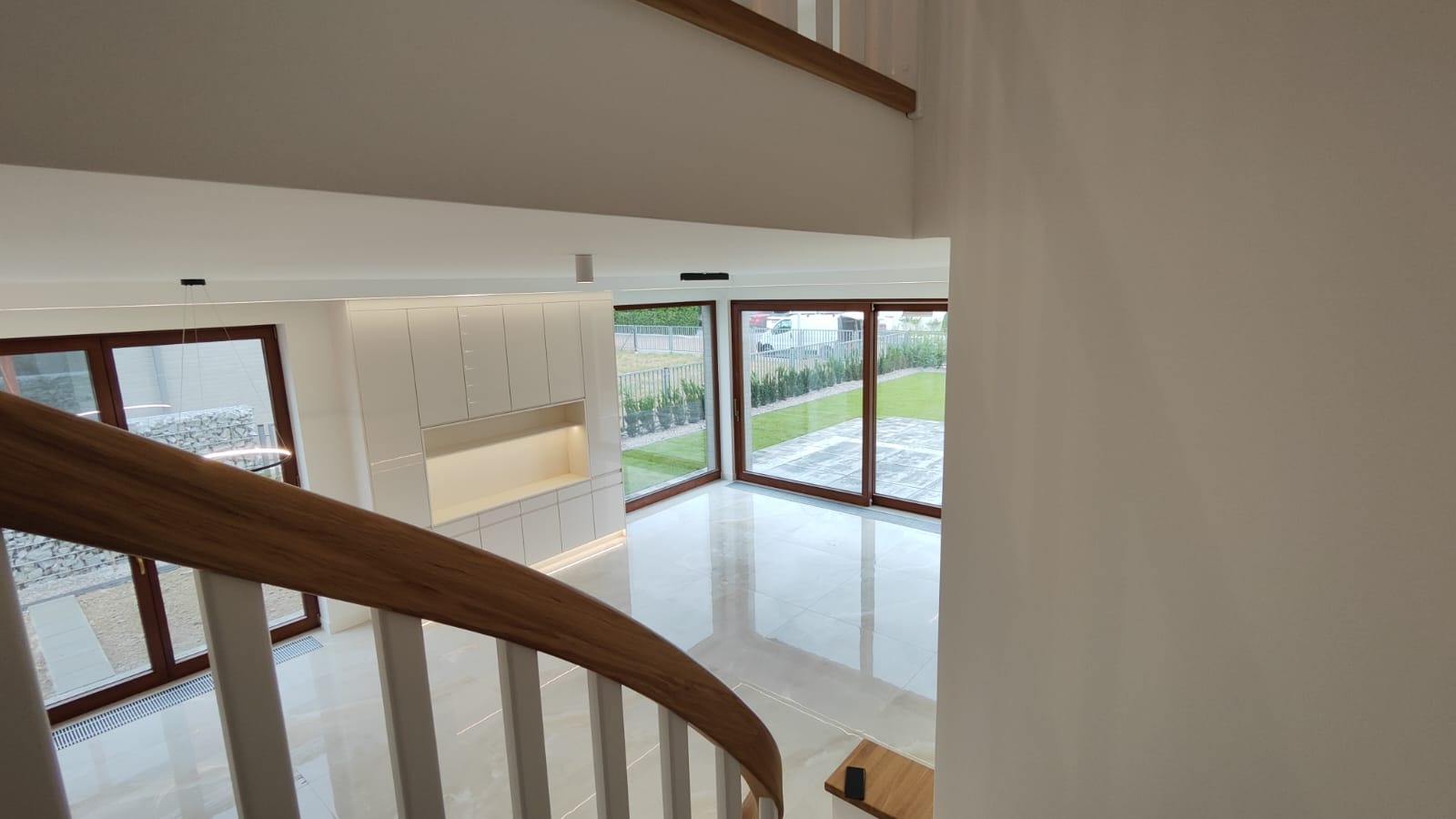 Nowoczesne wnętrze domu z widokiem na ogród, jasne płytki podłogowe, drewniane okna, białe ściany i zabudowa meblowa, widok z góry z perspektywy schodów.