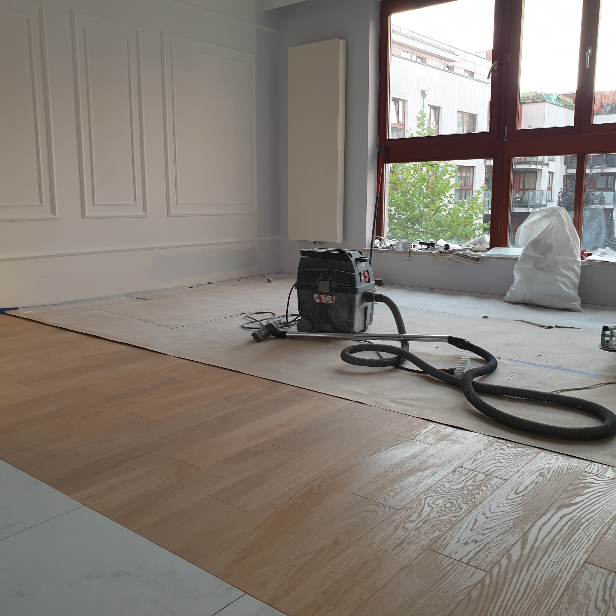 Układanie jasnych paneli podłogowych w pokoju z białymi ścianami i dużym oknem. Widoczny odkurzacz przemysłowy i zabezpieczona podłoga.