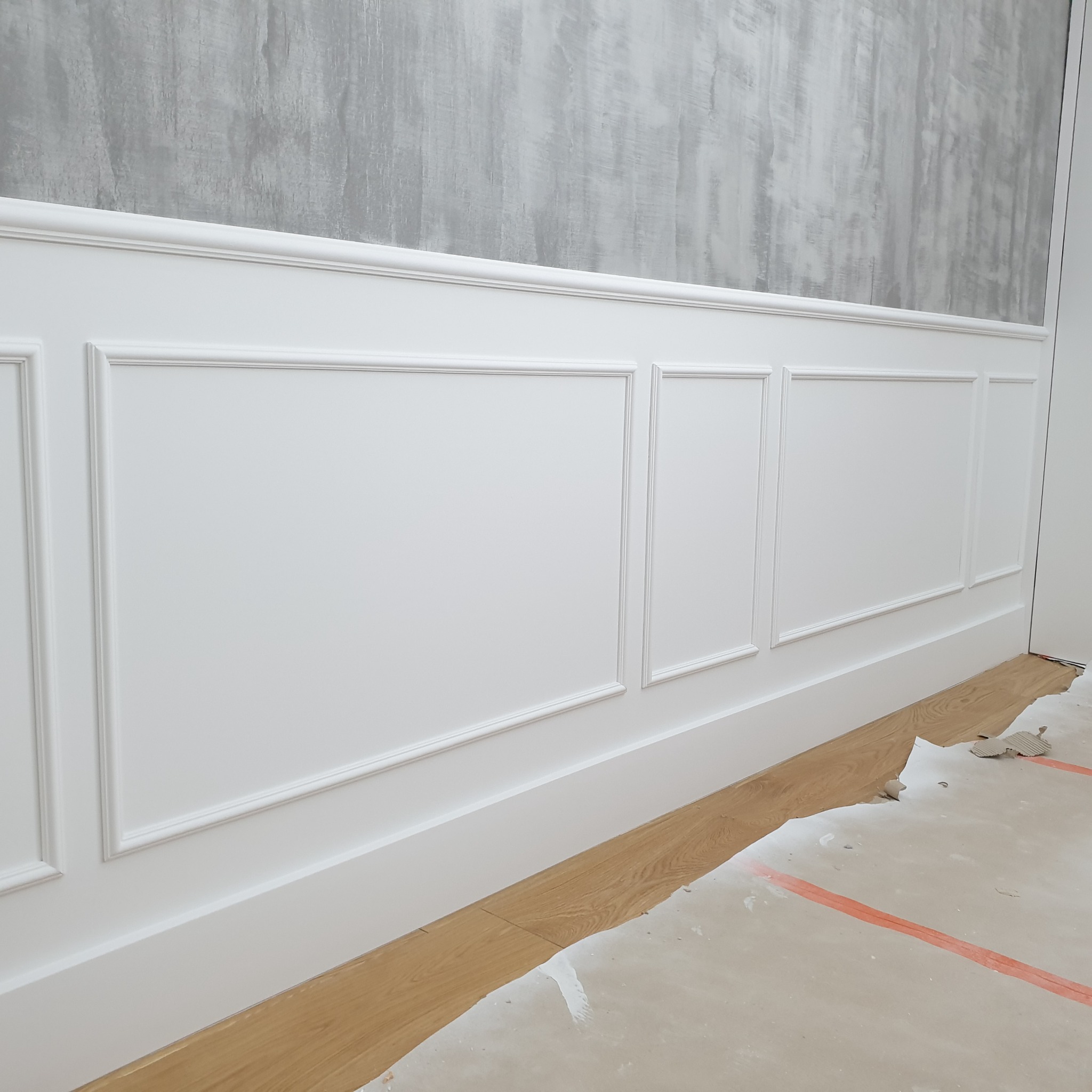 Biała boazeria angielska na ścianie z szarą, dekoracyjną farbą. Widoczne fragmenty podłogi z panelami i resztki po remoncie. Detal wykończenia wnętrza.