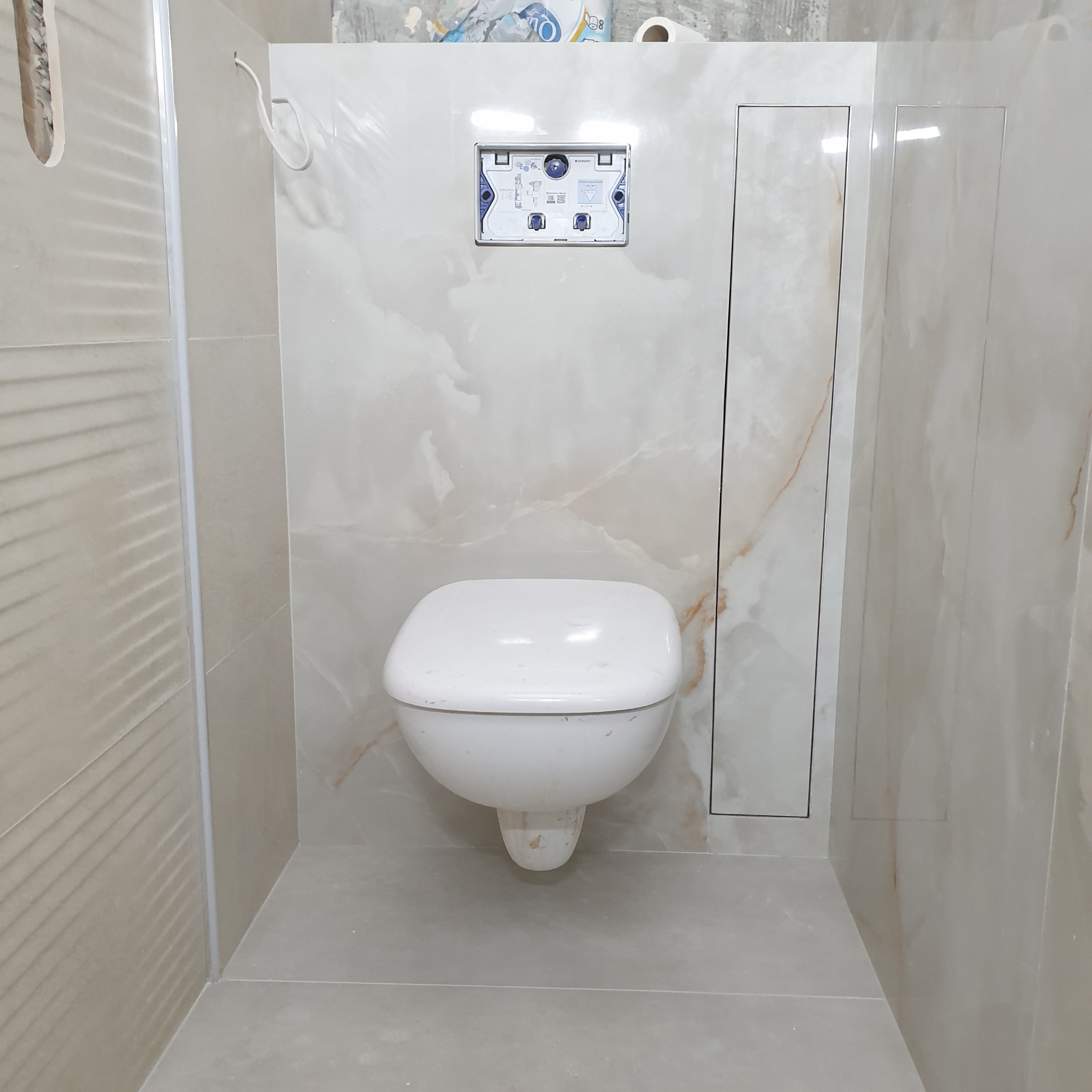 Wykończona toaleta podwieszana z beżowymi płytkami imitującymi marmur, widoczny stelaż podtynkowy i rewizja. Minimalistyczny design, ciepłe oświetlenie.