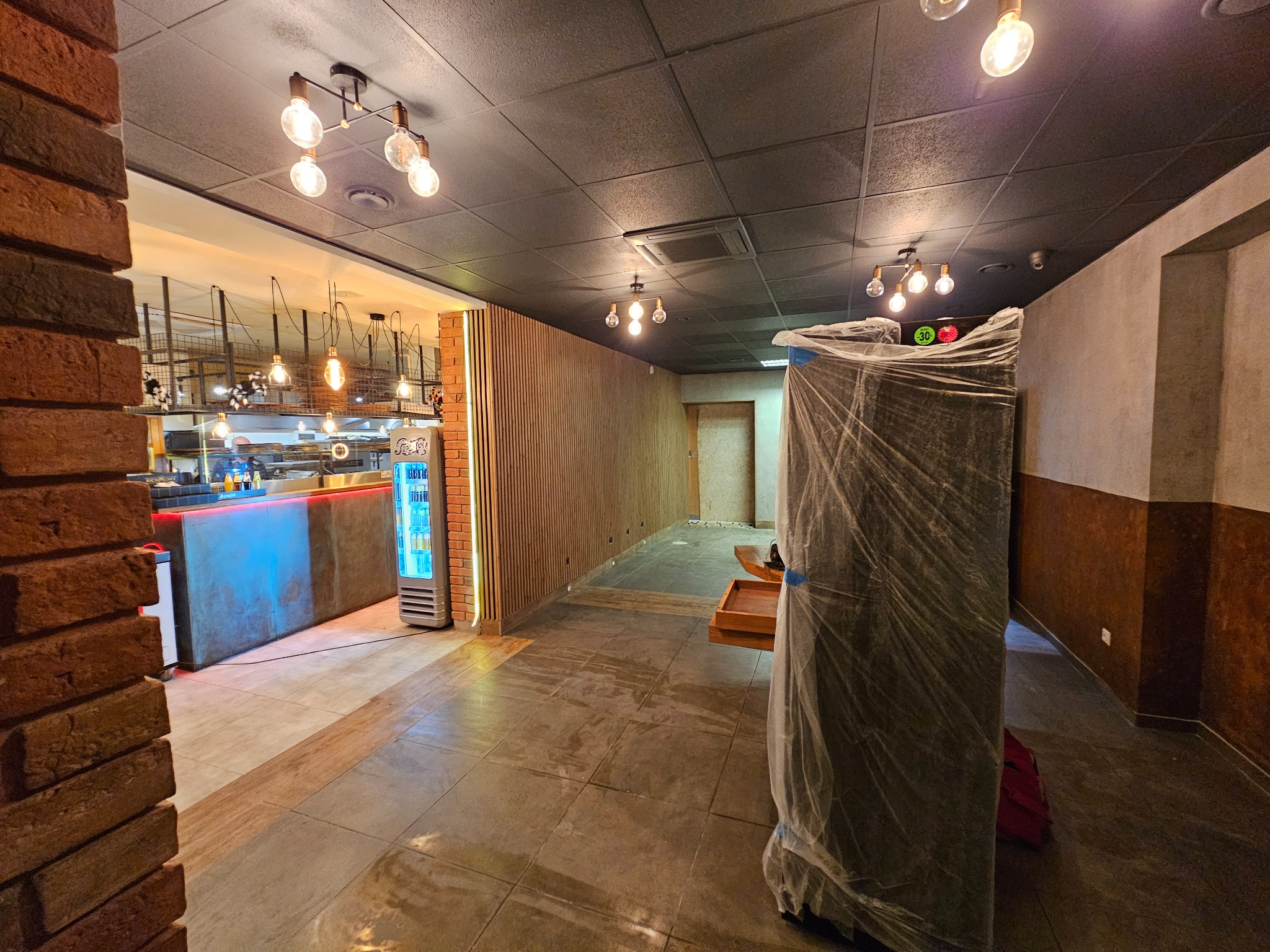 Wnętrze lokalu gastronomicznego w trakcie wykończenia: sufit z kasetonów, ceglana ściana, drewniane lamele, posadzka z płytek. Meble zabezpieczone folią.