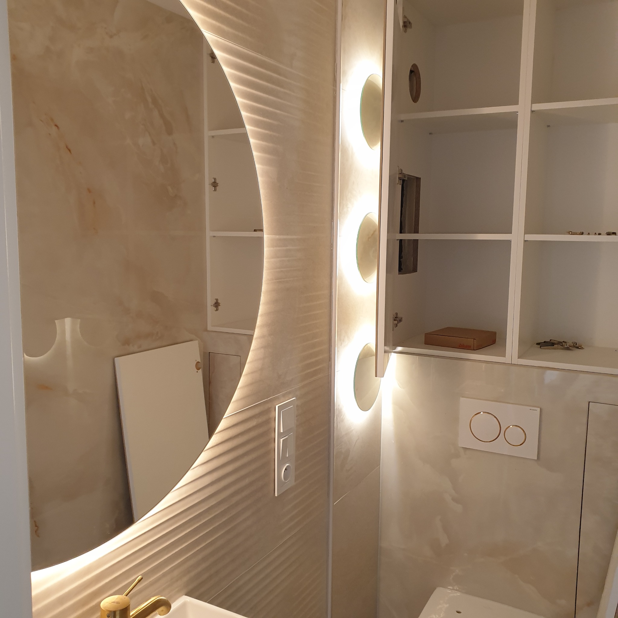 Wykończona łazienka z okrągłym lustrem z podświetleniem LED, szafką w zabudowie i płytkami imitującymi marmur. Złota bateria umywalkowa i biała armatura.