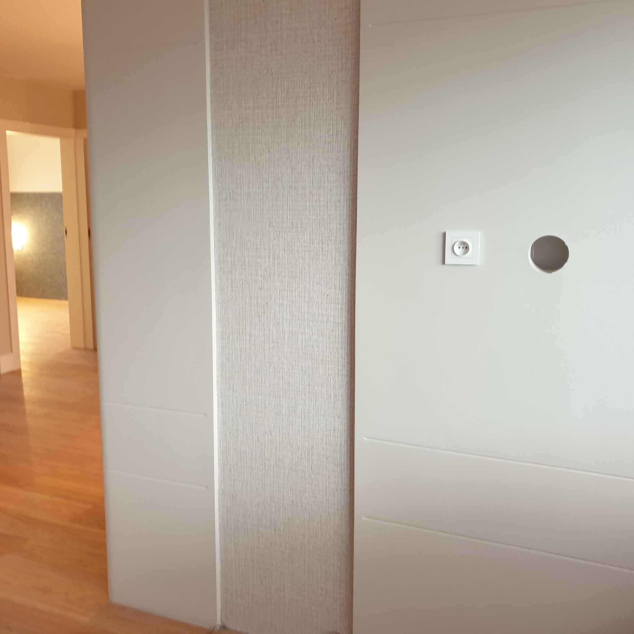 Ściana po remoncie z tapetą strukturalną, gniazdkiem i otworem na kabel. Widoczna podłoga z paneli i fragment innego pomieszczenia w tle. Jasne, neutralne kolory.