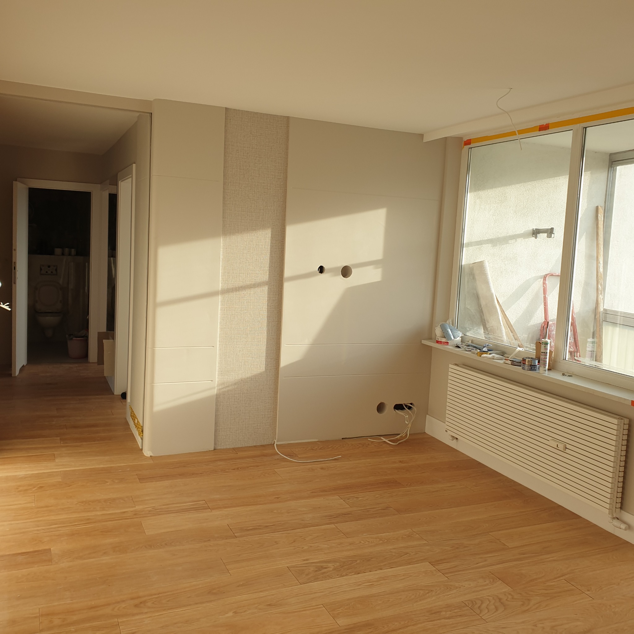 Jasne panele podłogowe w pokoju po remoncie. Ściana z dekoracyjną tapetą i przygotowanymi otworami na gniazdka. Widoczne narzędzia na parapecie. W tle łazienka.