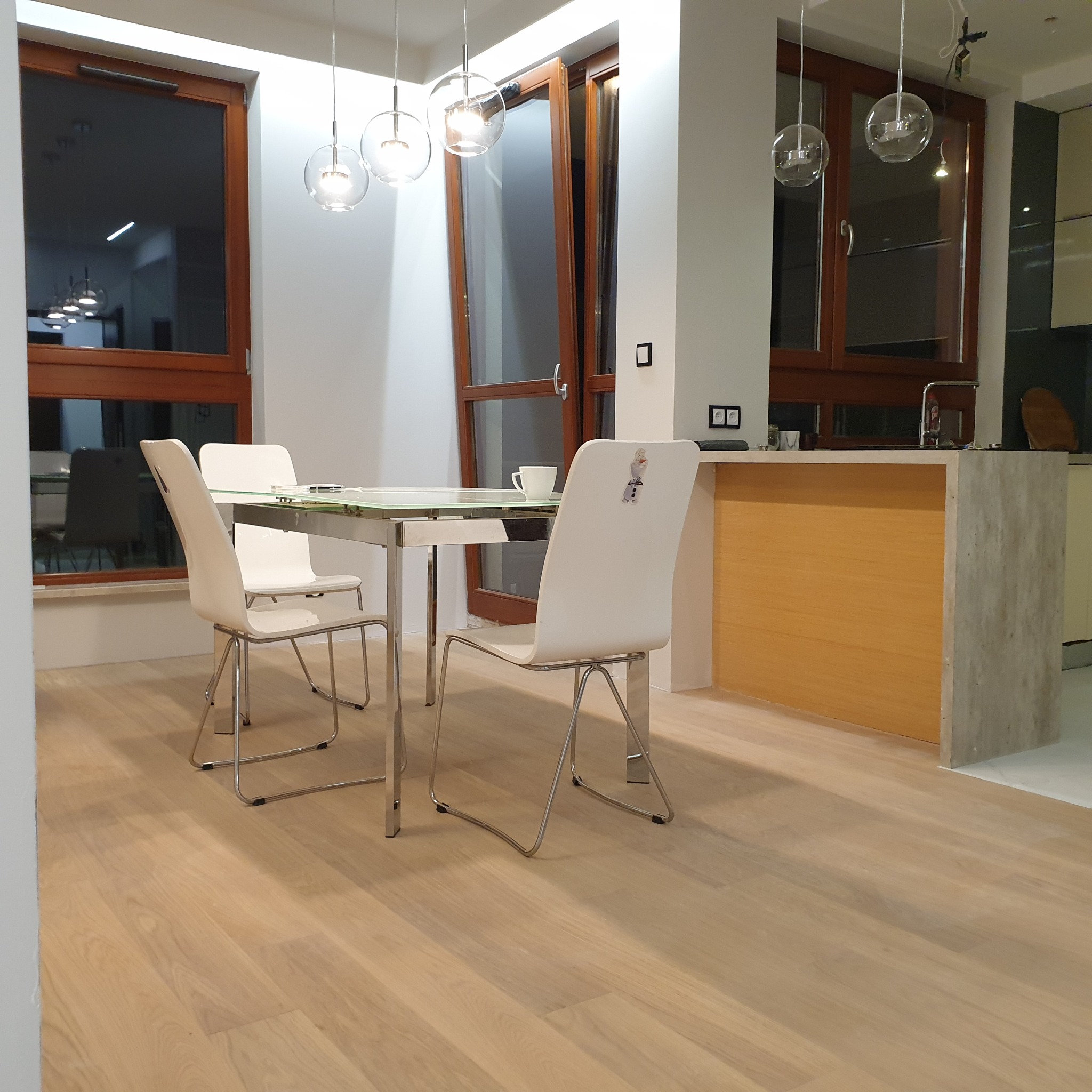 Jasna, nowoczesna jadalnia z drewnianą podłogą, szklanym stołem i białymi krzesłami. Widoczne okna z drewnianą ramą i lampy wiszące w kształcie kul.