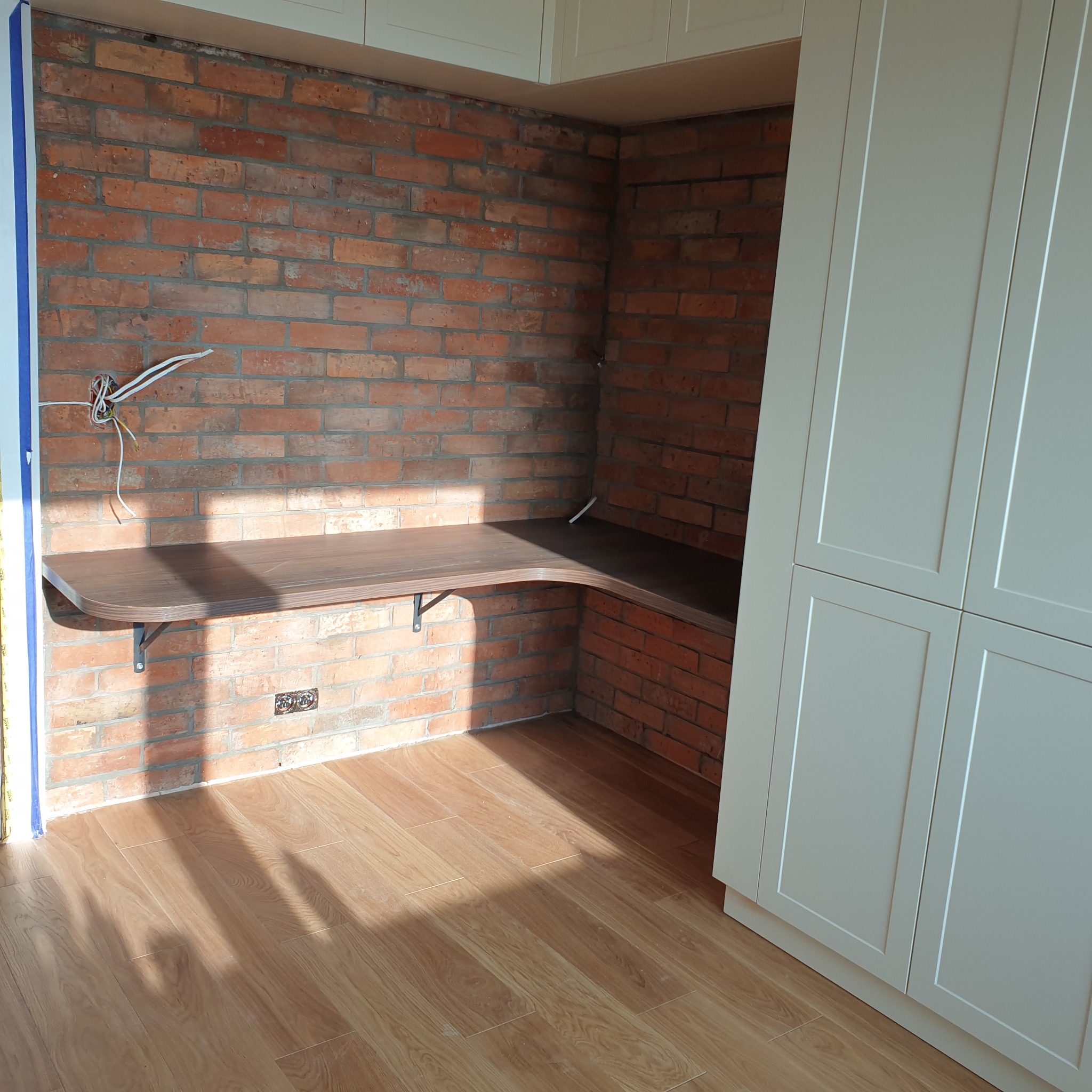 Kącik z biurkiem z blatem w kolorze drewna, wspartym na metalowych wspornikach, na tle ściany z czerwonej cegły i białej zabudowy meblowej. Widoczne przewody elektryczne.