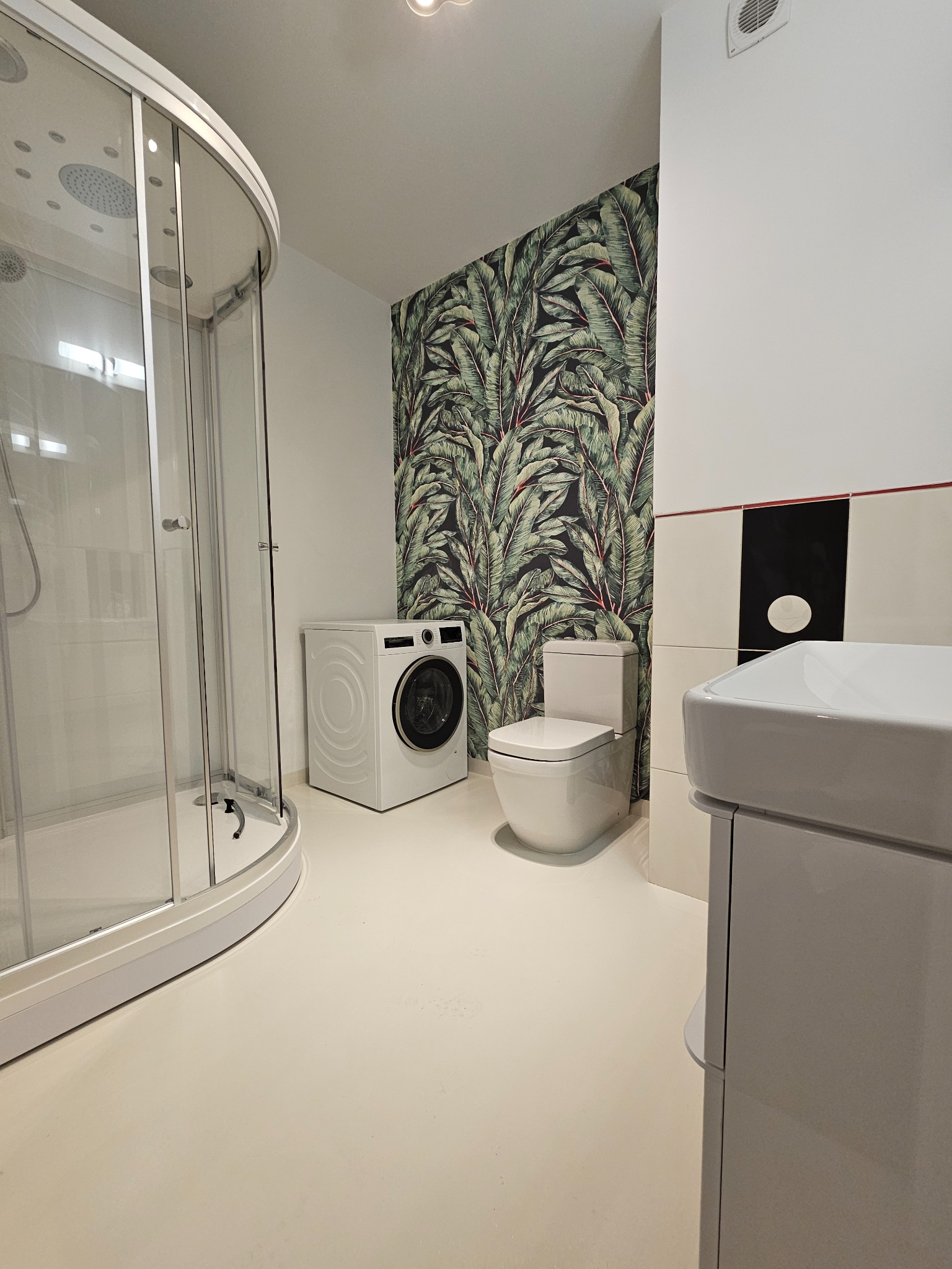 Nowoczesna łazienka z kabiną prysznicową, pralką i toaletą na tle ściany z motywem liści. Minimalistyczny design, jasne kolory, oświetlenie sufitowe.