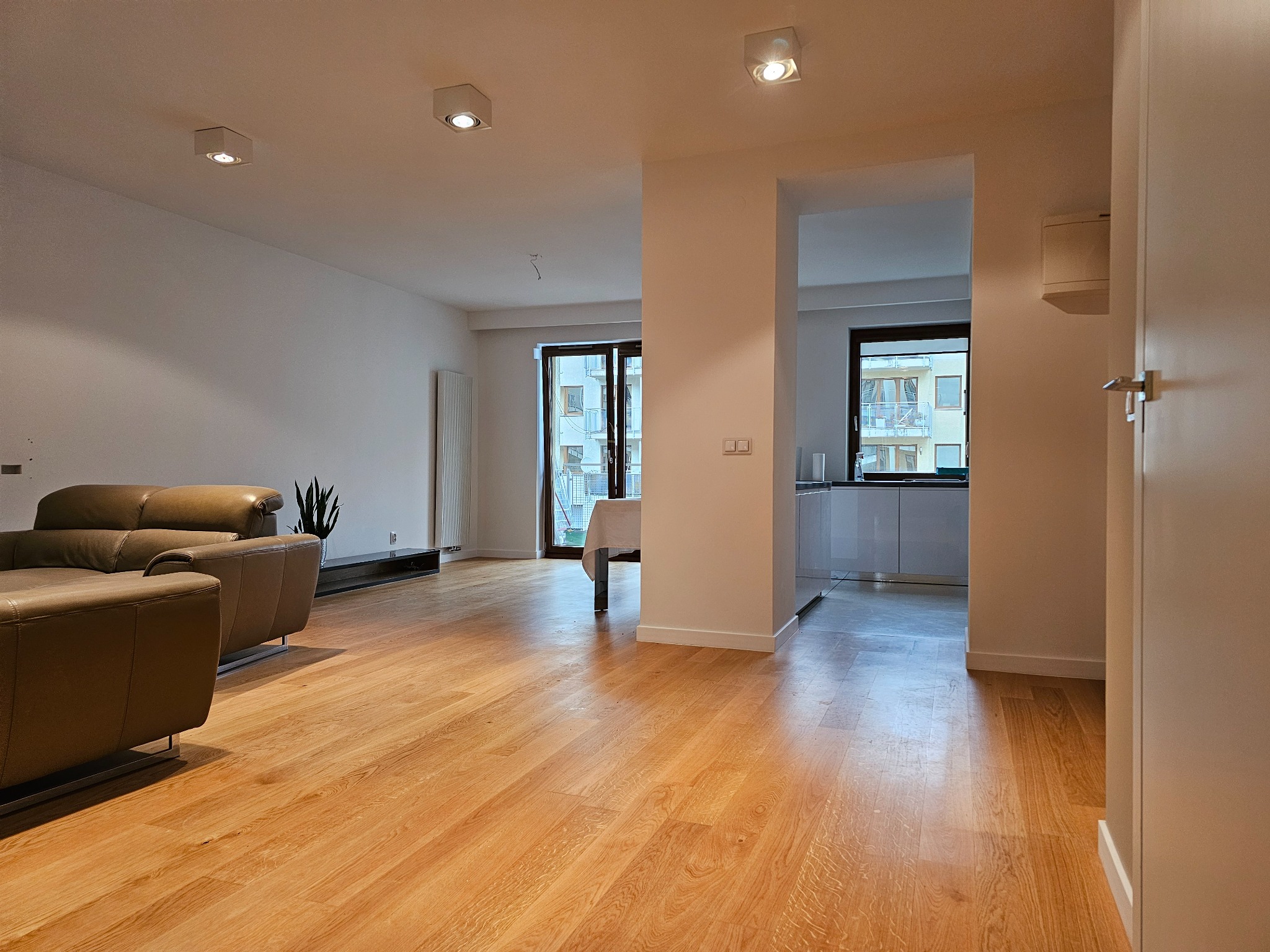 Jasne panele podłogowe w przestronnym salonie z beżową sofą i widokiem na kuchnię. Minimalistyczne oświetlenie sufitowe i białe ściany. Widok na zewnątrz przez okno.