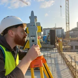Mężczyzna w kasku i kamizelce odblaskowej obsługuje niwelator optyczny GeoMax Zoom 80 na budowie, w tle widoczny dźwig i powstający budynek.
