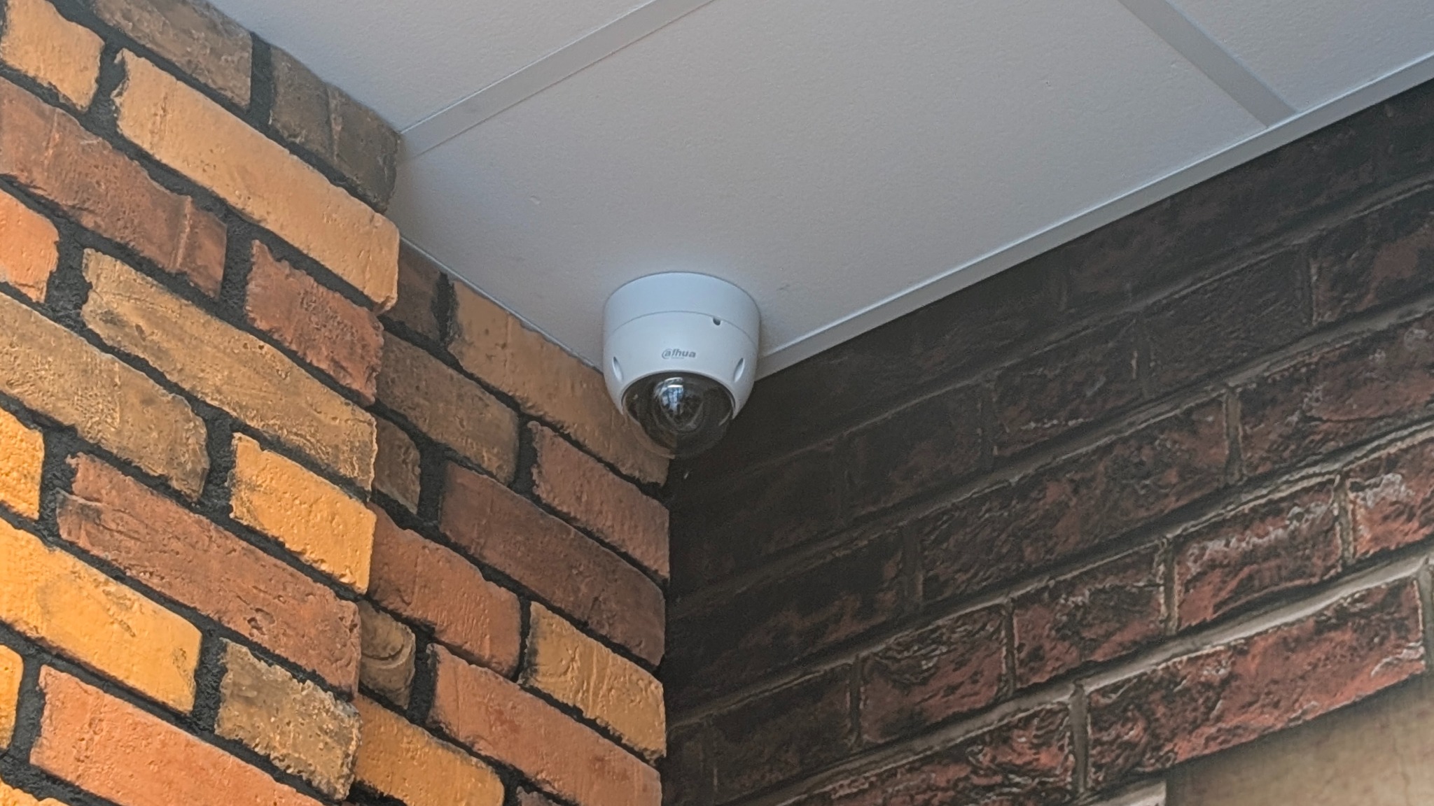 Kamera kopułkowa Dahua zamontowana w rogu pomieszczenia, na styku białego sufitu i ściany z cegły w odcieniach brązu i pomarańczy. Widok z dołu, lekko z ukosa.