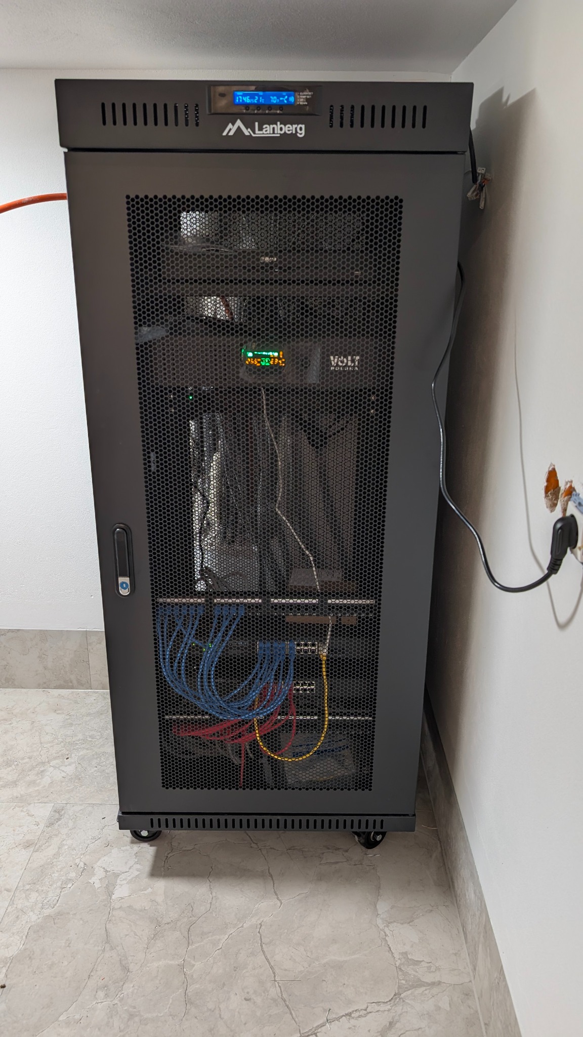 Czarna szafa rack Lanberg z zainstalowanym sprzętem sieciowym (kable, switche) w pomieszczeniu z marmurową podłogą. Widoczne okablowanie i zasilanie.