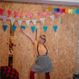 Osoba w okularach i paski ubraniu, z gwizdkiem w ustach, tańczy na tle ściany z dekoracjami imprezowymi, w tym kolorowe girlandy i lampki.