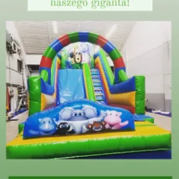 Duży, kolorowy dmuchany zamek ze zjeżdżalnią, ozdobiony motywami zwierząt, ustawiony w hali.