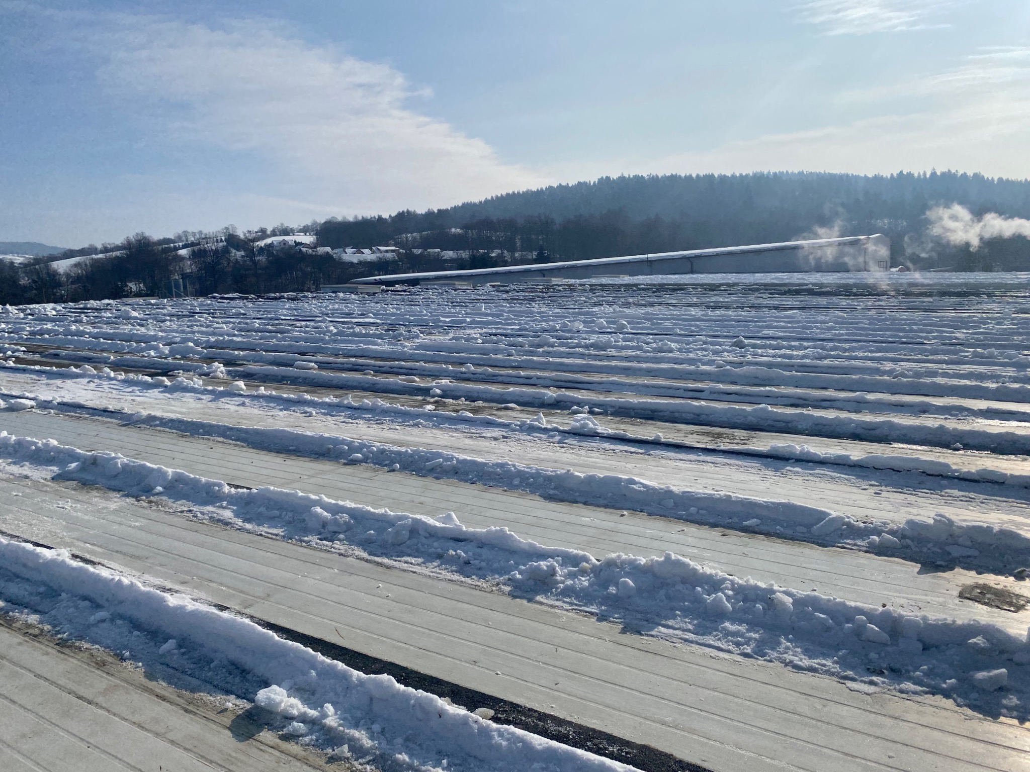 Śnieg zalegający na płaskim dachu z widocznymi pasami odśnieżonymi, w tle zalesione wzgórza i budynek z komina którego wydobywa się para.