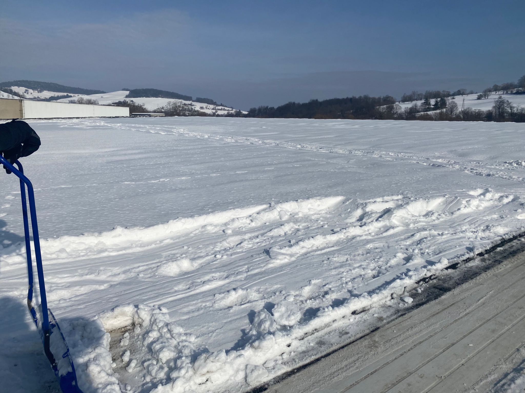 Błękitna łopata do śniegu częściowo widoczna na tle rozległego, białego dachu pokrytego śniegiem z odśnieżonymi pasami, w oddali zalesione wzgórza i zabudowania w pochmurny dzień.