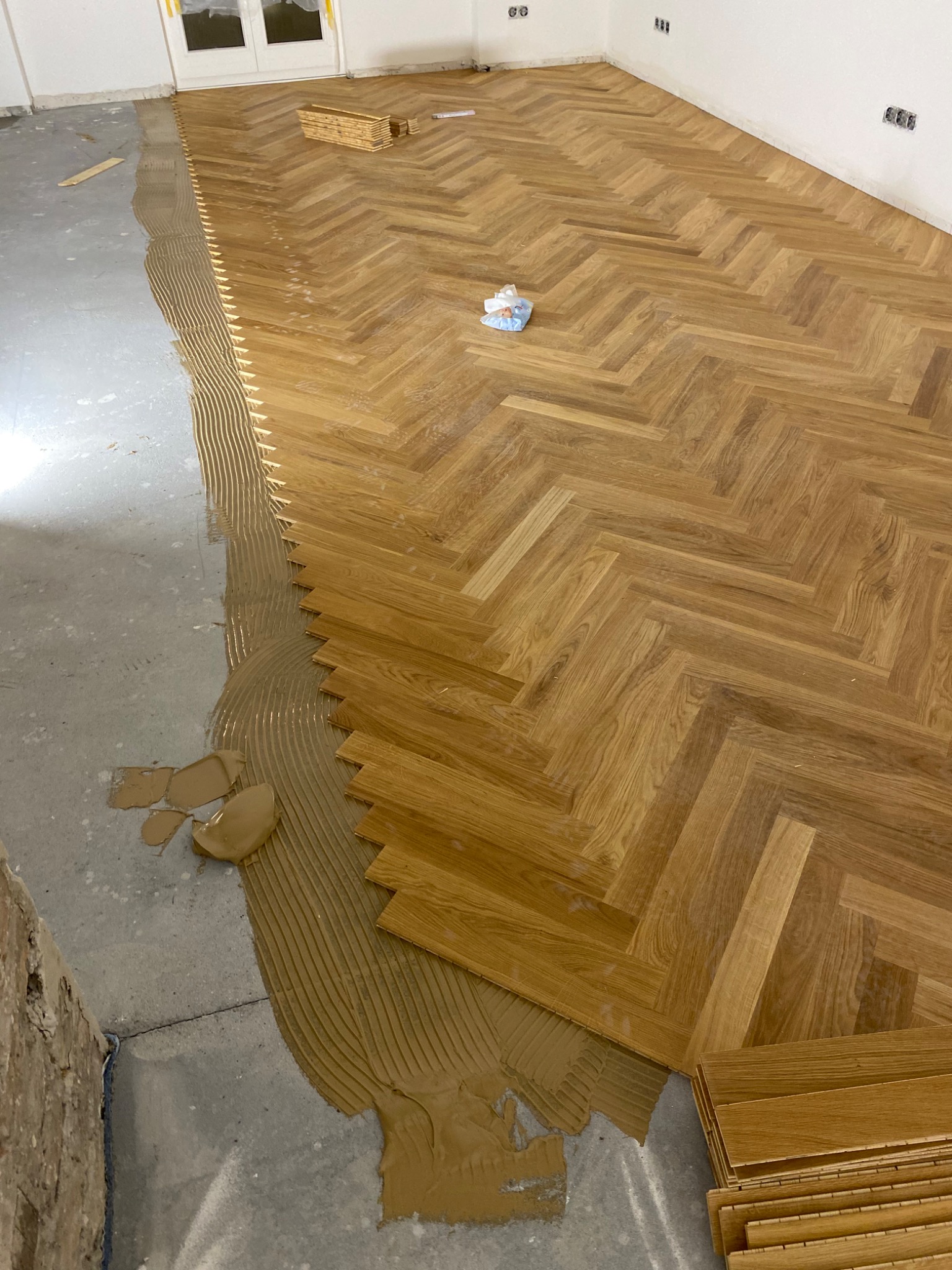 Montaż parkietu w jodełkę w Krosno: widok na klej rozprowadzony na podłodze i ułożoną część parkietu z dębowego drewna, z paczką desek gotowych do ułożenia.