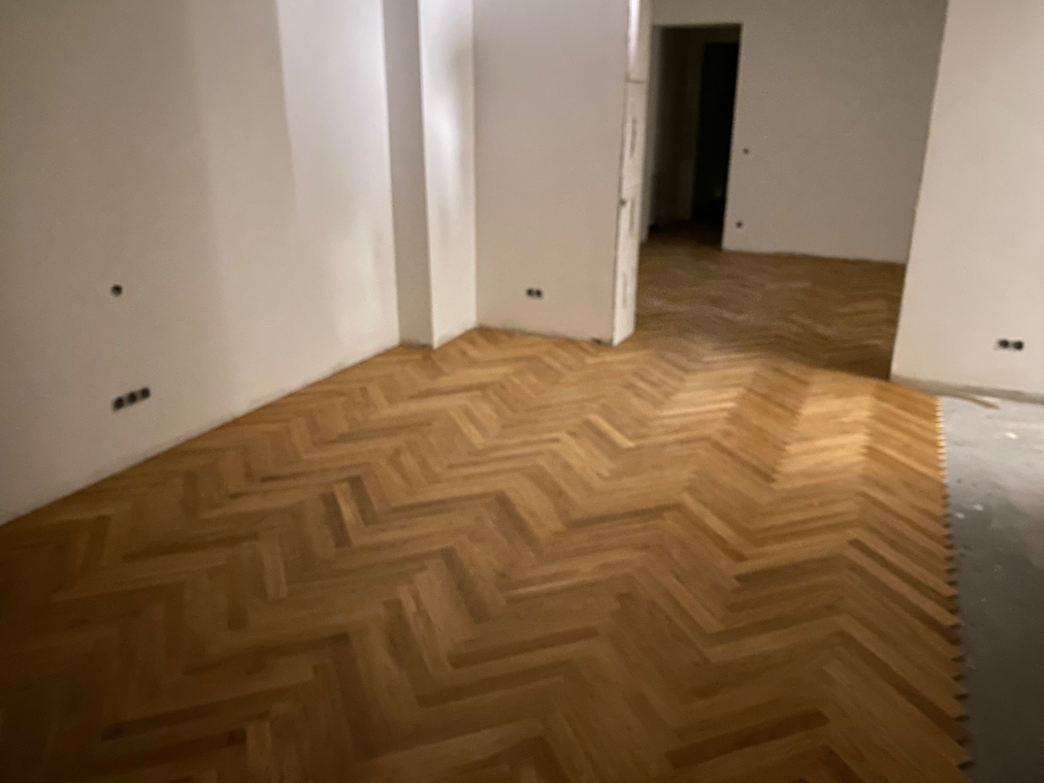 Nowo ułożony parkiet w jodełkę w pustym pokoju z białymi ścianami i widocznymi gniazdkami elektrycznymi, z otwartym przejściem do kolejnego pomieszczenia.