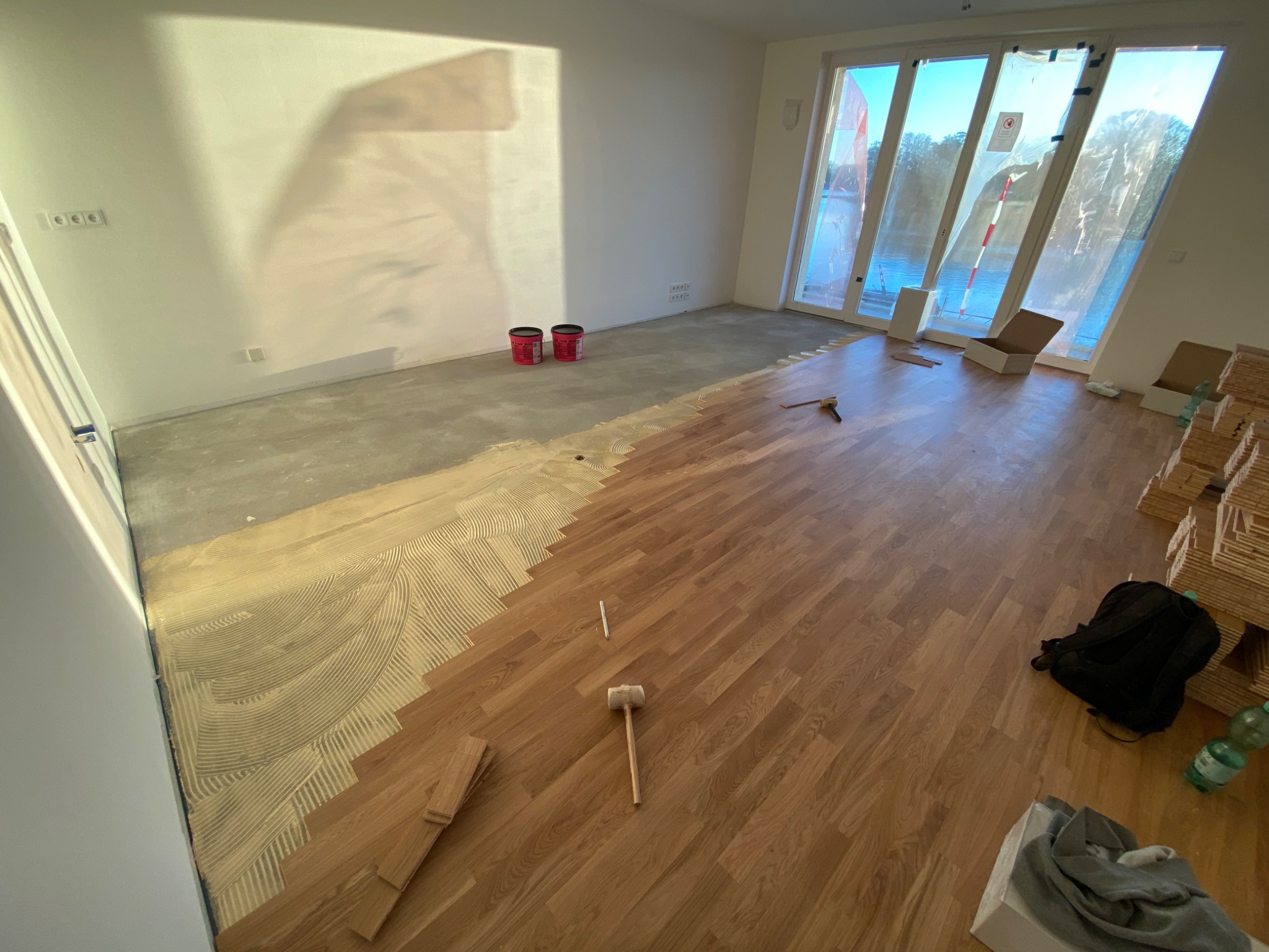 Proces montażu drewnianej podłogi: widoczny klej na podłożu, ułożone deski, gumowy młotek, zapas desek i okna z widokiem na zewnątrz.