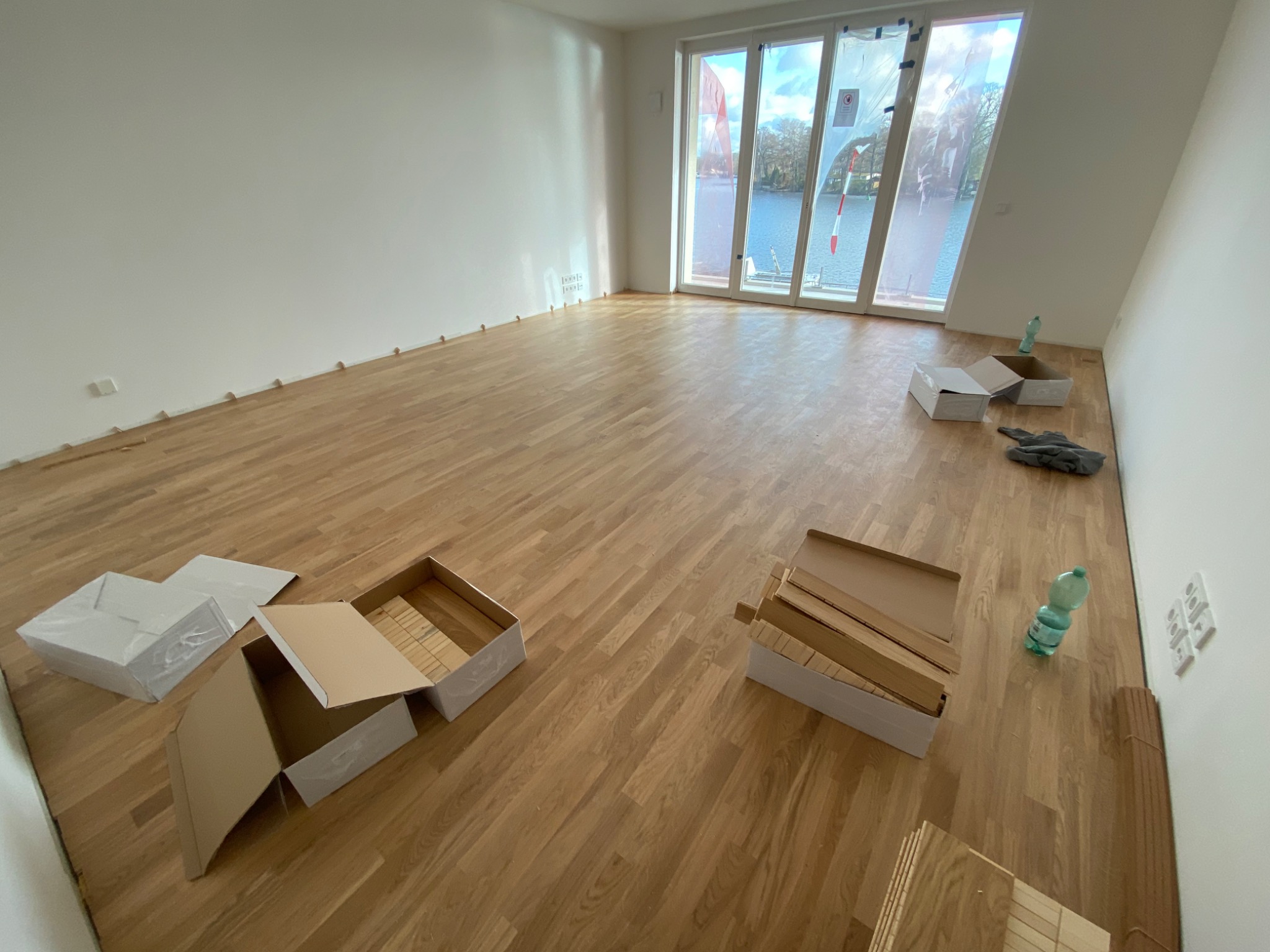 Wnętrze pokoju z drewnianą podłogą w trakcie montażu, widoczne otwarte kartony z panelami podłogowymi, listwy przypodłogowe i widok na jezioro za oknem.