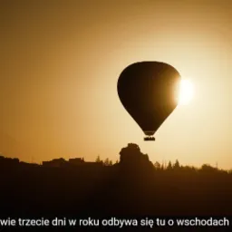 Sylwetka balonu na tle wschodzącego słońca, zarys gór i zabudowań w oddali, ciepła, złota kolorystyka.