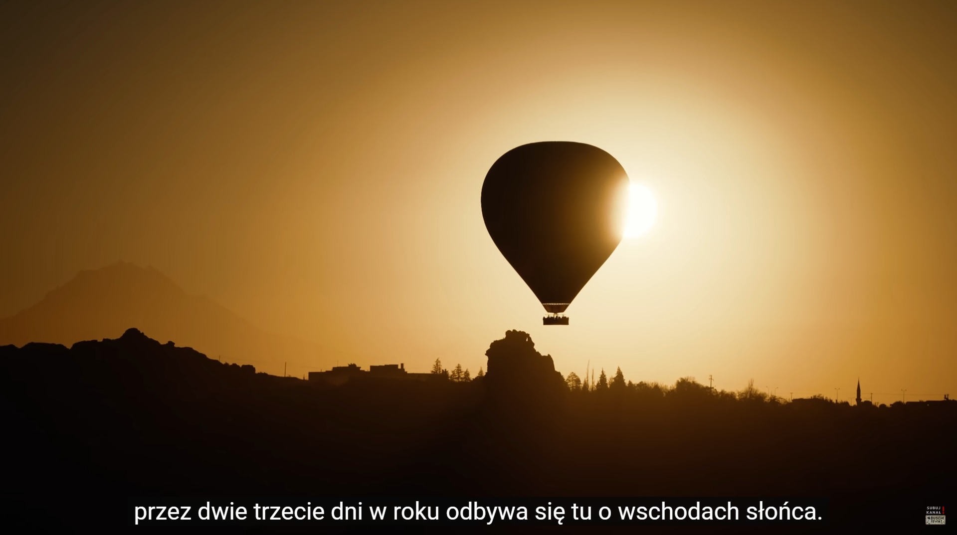 Sylwetka balonu na tle wschodzącego słońca, zarys gór i zabudowań w oddali, ciepła, złota kolorystyka.