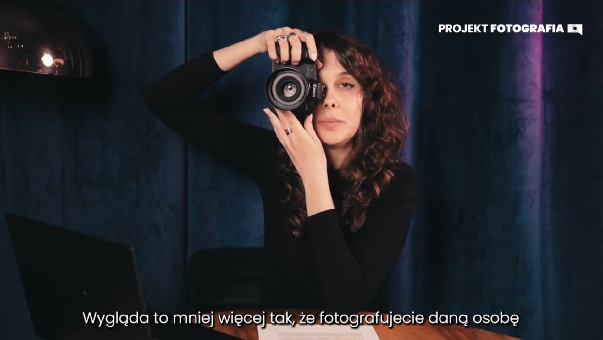 Kobieta z kręconymi włosami trzyma aparat Canon EOS w dłoniach, spoglądając w obiektyw, widoczny laptop i ciemnoniebieskie tło.