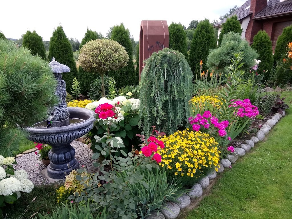 Ogród z różnorodną roślinnością, fontanną z rzeźbą i obrzeżem z kamieni polnych, z tujami w tle i elementem architektury ogrodowej.