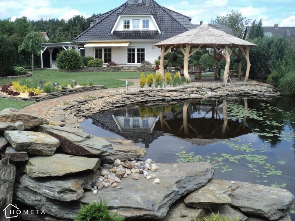 Oczko wodne z kamiennym brzegiem i liliami wodnymi, altana z drewnianymi słupami odbijająca się w wodzie, ścieżka z kamieni prowadząca do domu z ogrodem.