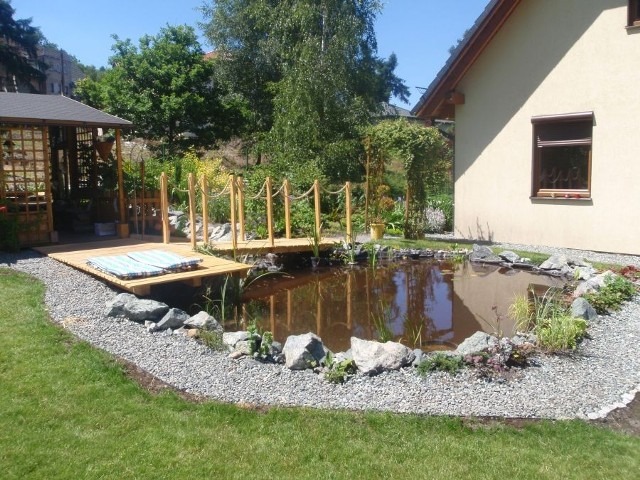 Oczko wodne z drewnianym pomostem i mostkiem z linowymi barierkami, otoczone kamieniami, z widoczną altaną w tle i zielenią ogrodową.