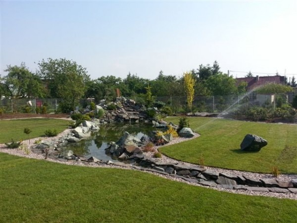 Ogród z oczkiem wodnym i kaskadą skalną, otoczony trawnikiem, z systemem nawadniania w tle.