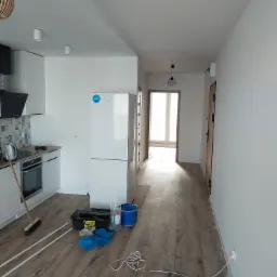 Wnętrze mieszkania w trakcie remontu: panele podłogowe ułożone, widoczne narzędzia, listwy przypodłogowe, kabel zasilający na podłodze, kuchnia z białymi szafkami i lodówką w tle.