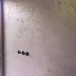 Ściana z dekoracyjnym tynkiem w odcieniach beżu i brązu, z widocznymi puszkami elektrycznymi przed montażem gniazdek.