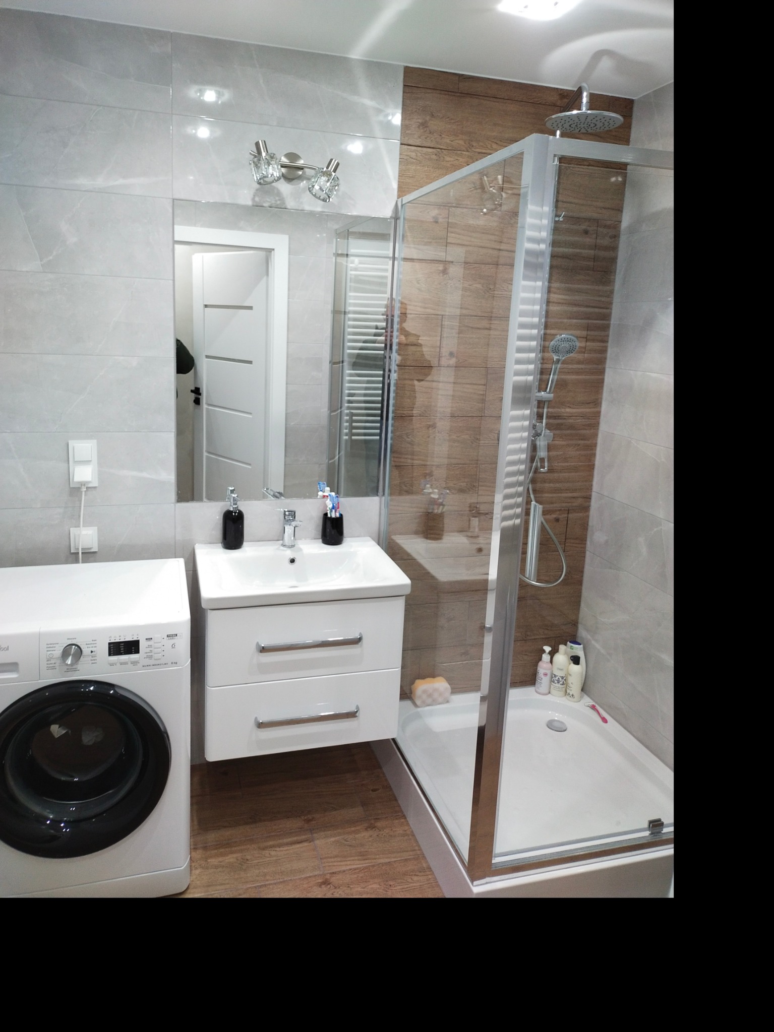 Nowoczesna łazienka z szarymi płytkami imitującymi marmur, drewnopodobnymi akcentami w kabinie prysznicowej, białą umywalką z szafką, pralką i lustrem z oświetleniem.