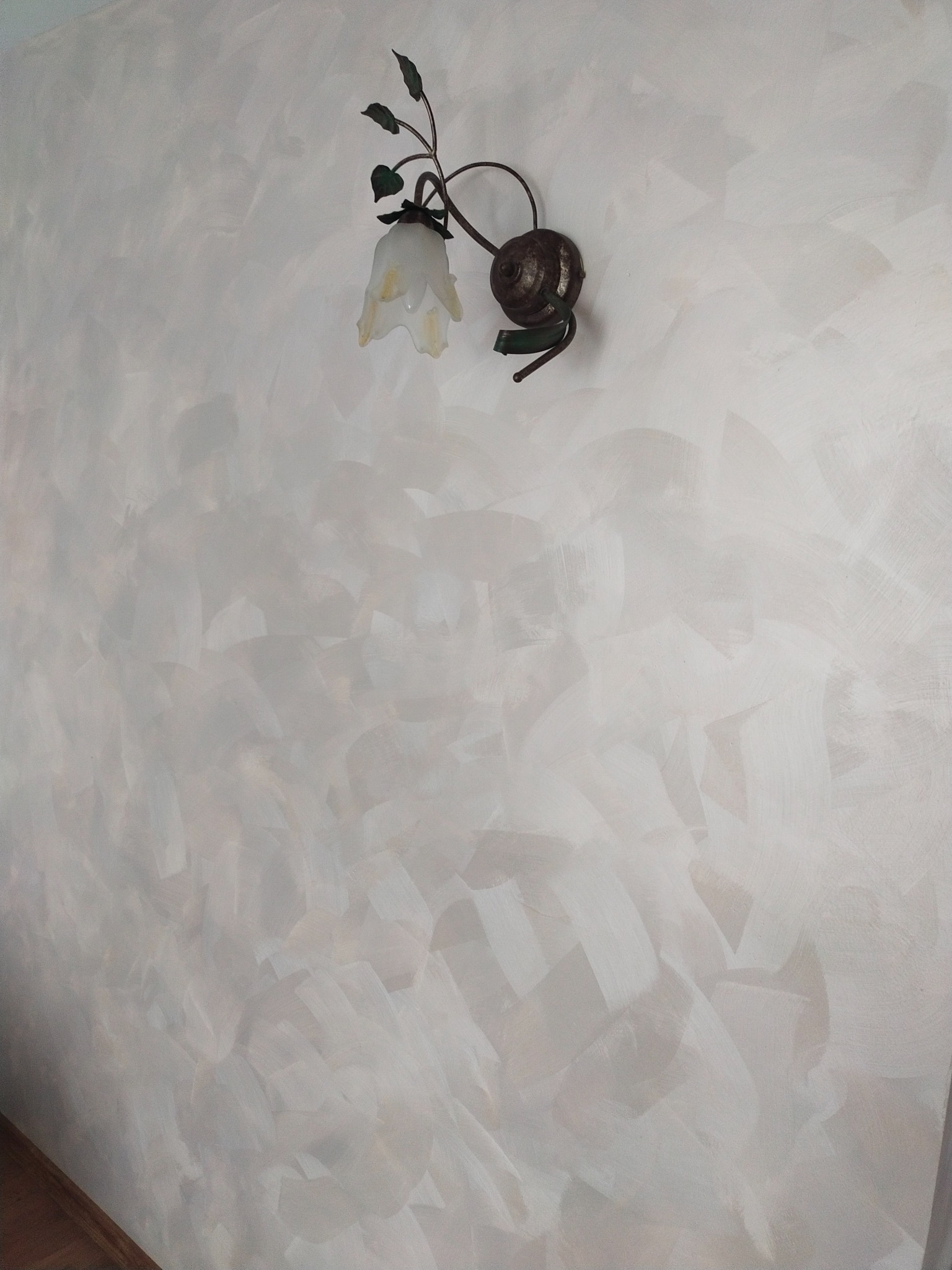 Ściana z dekoracyjną, strukturalną farbą w odcieniach bieli i szarości, oświetlona przez kinkiet w stylu retro z kwiatowym kloszem.