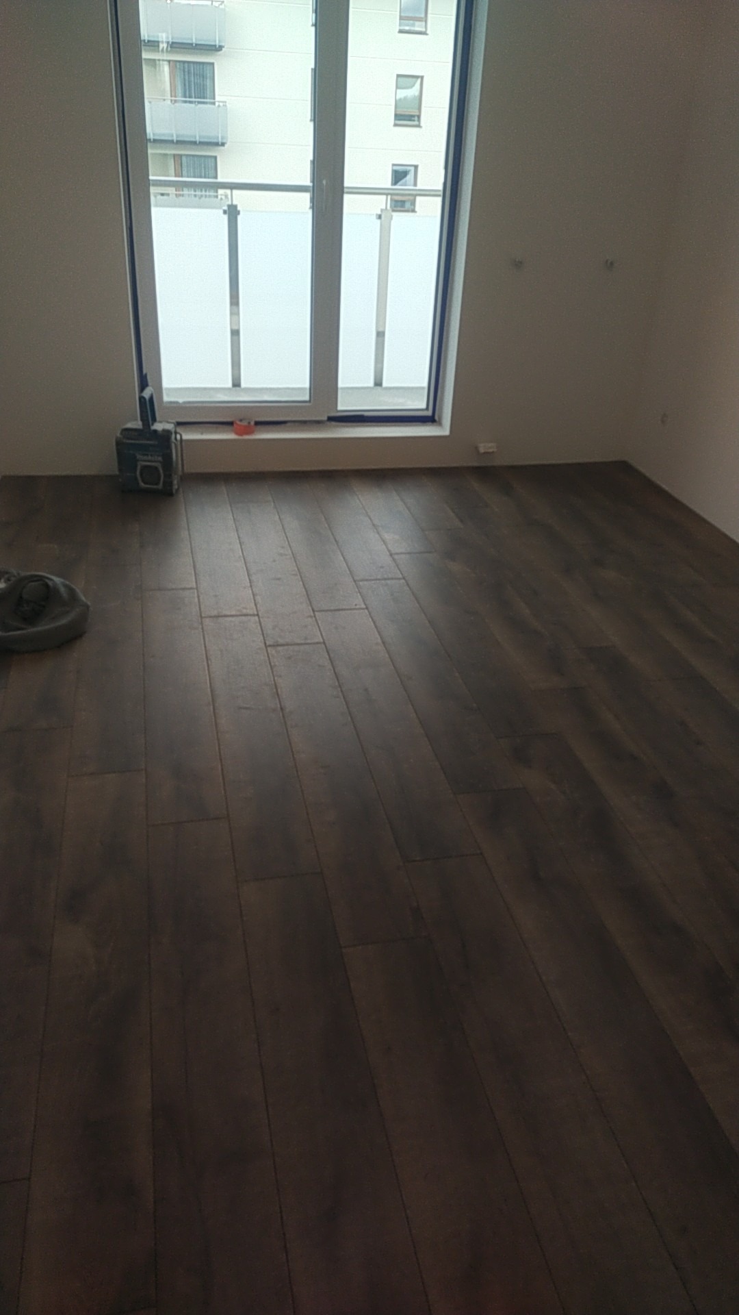Ułożone, ciemne panele podłogowe w pustym pokoju z dużym oknem balkonowym, widoczny sprzęt do montażu paneli.