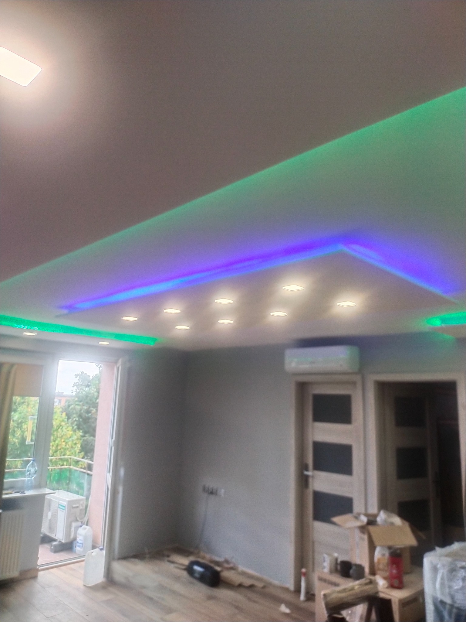 Nowoczesny sufit podwieszany z oświetleniem LED w kolorach zielonym i fioletowym, z widocznymi punktami świetlnymi, klimatyzator na ścianie, oraz częściowo widoczne drzwi z dekoracyjnymi panelami...