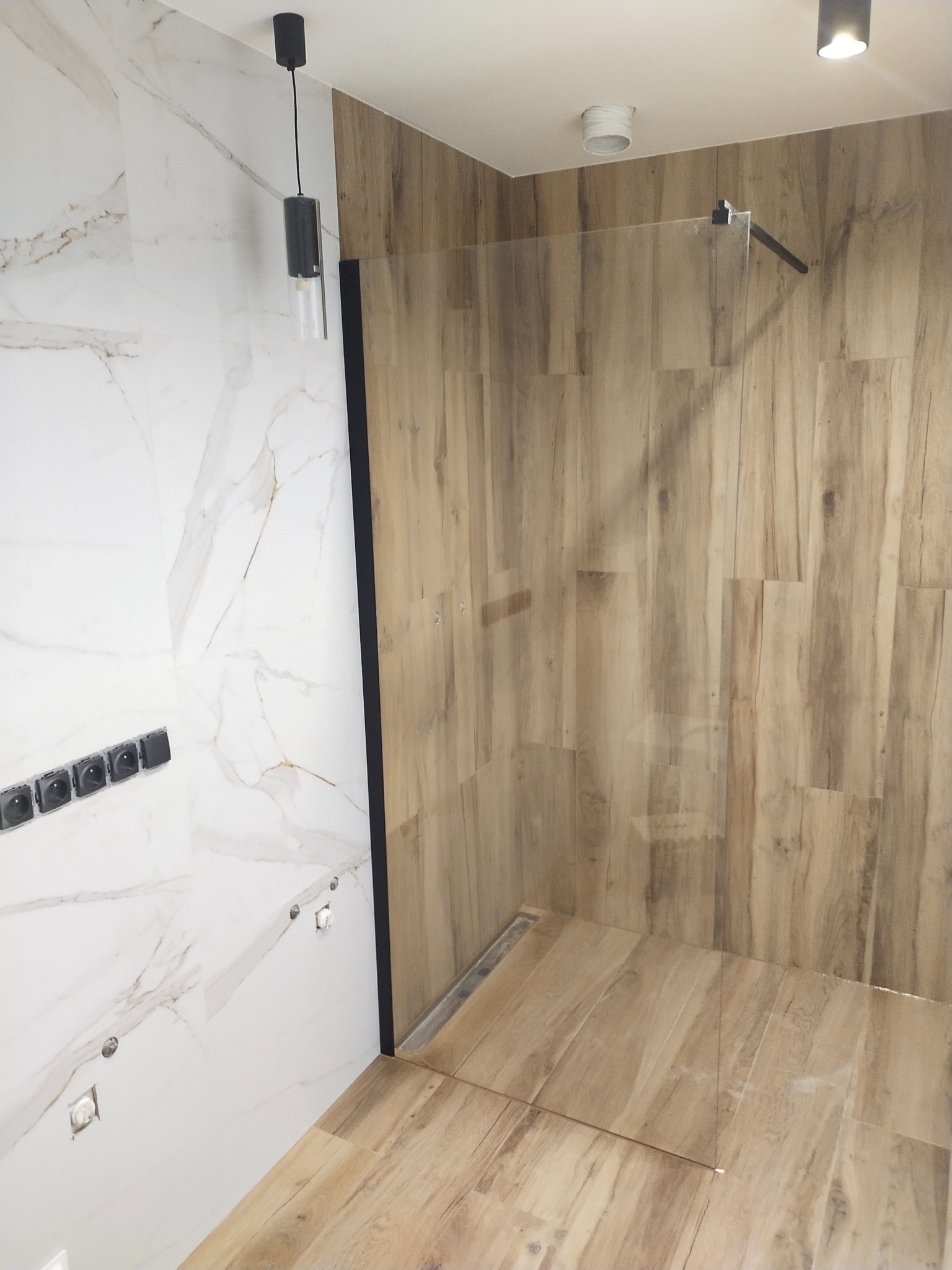 Nowoczesna kabina prysznicowa walk-in z drewnopodobnymi płytkami na ścianach i podłodze, biała marmurowa ściana z czarnymi gniazdkami, minimalistyczna czarna lampa wisząca.