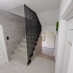 Nowoczesna, czarna barierka schodowa z pionowych prętów w domu w Porażu, na tle ściany z cegły i schodów wyłożonych jasnymi płytkami.