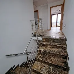 Schody z egzotycznego kamienia z nowoczesną, minimalistyczną barierką ze stali nierdzewnej. W tle widoczne drzwi do pomieszczeń z oknami.