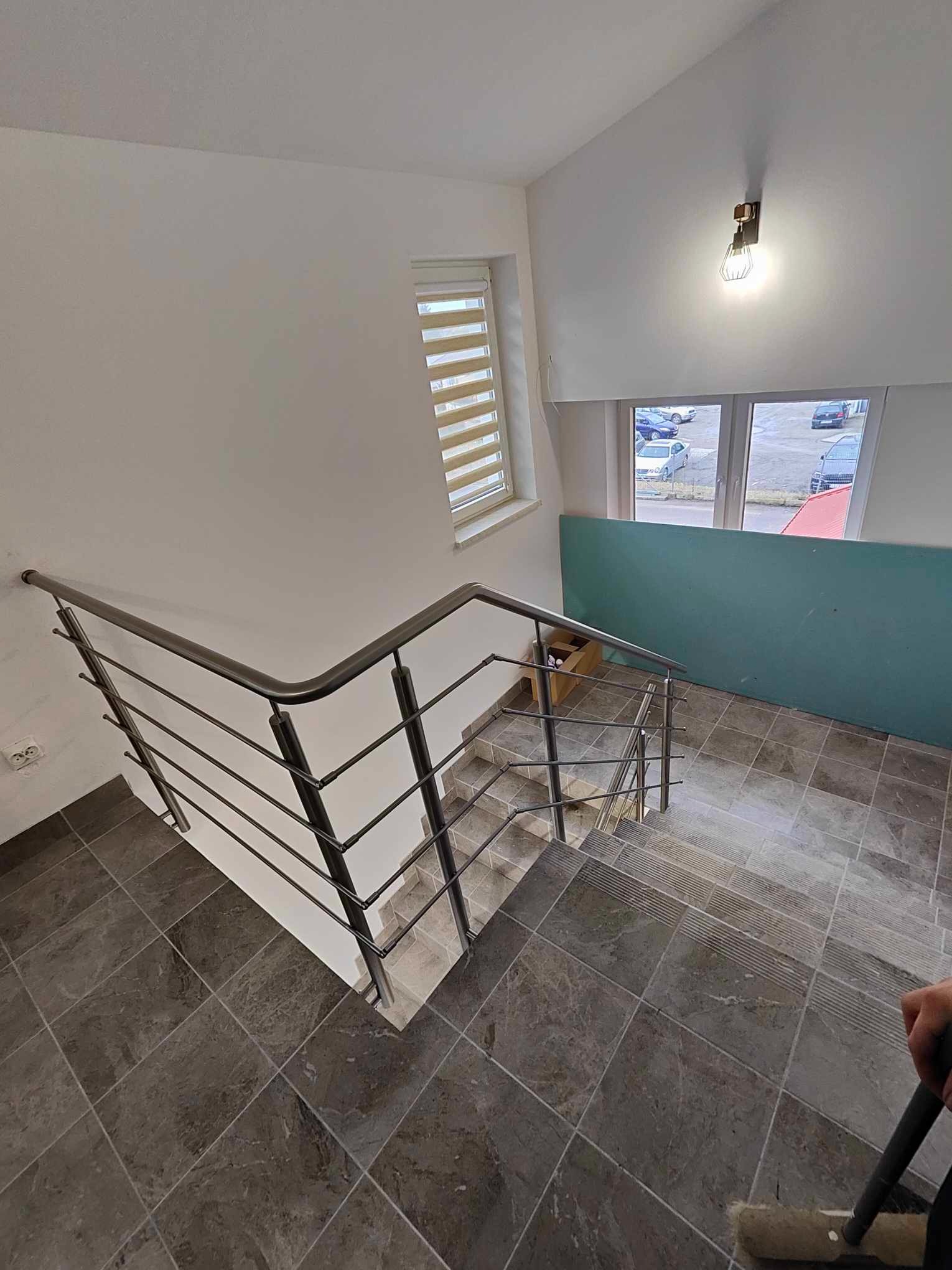 Wewnętrzna klatka schodowa z szarymi, marmurkowymi płytkami i balustradą ze stali nierdzewnej, widok z góry na dół, z oknem i żaluzjami w tle.