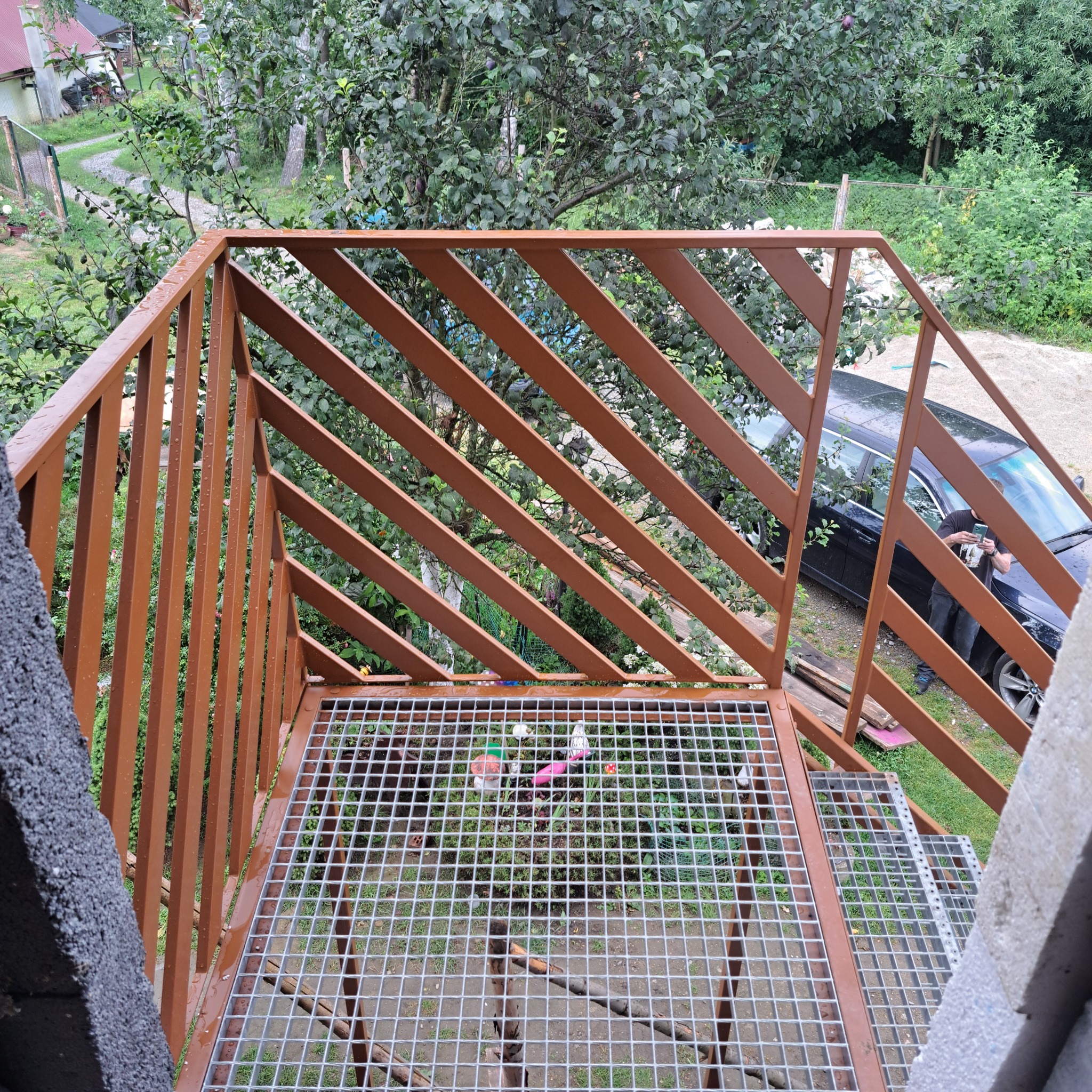 Brązowa metalowa konstrukcja schodów zewnętrznych z ukośnymi szczeblami, widok z góry na kratę podestu, w tle zieleń i czarny samochód z osobą robiącą zdjęcie telefonem.