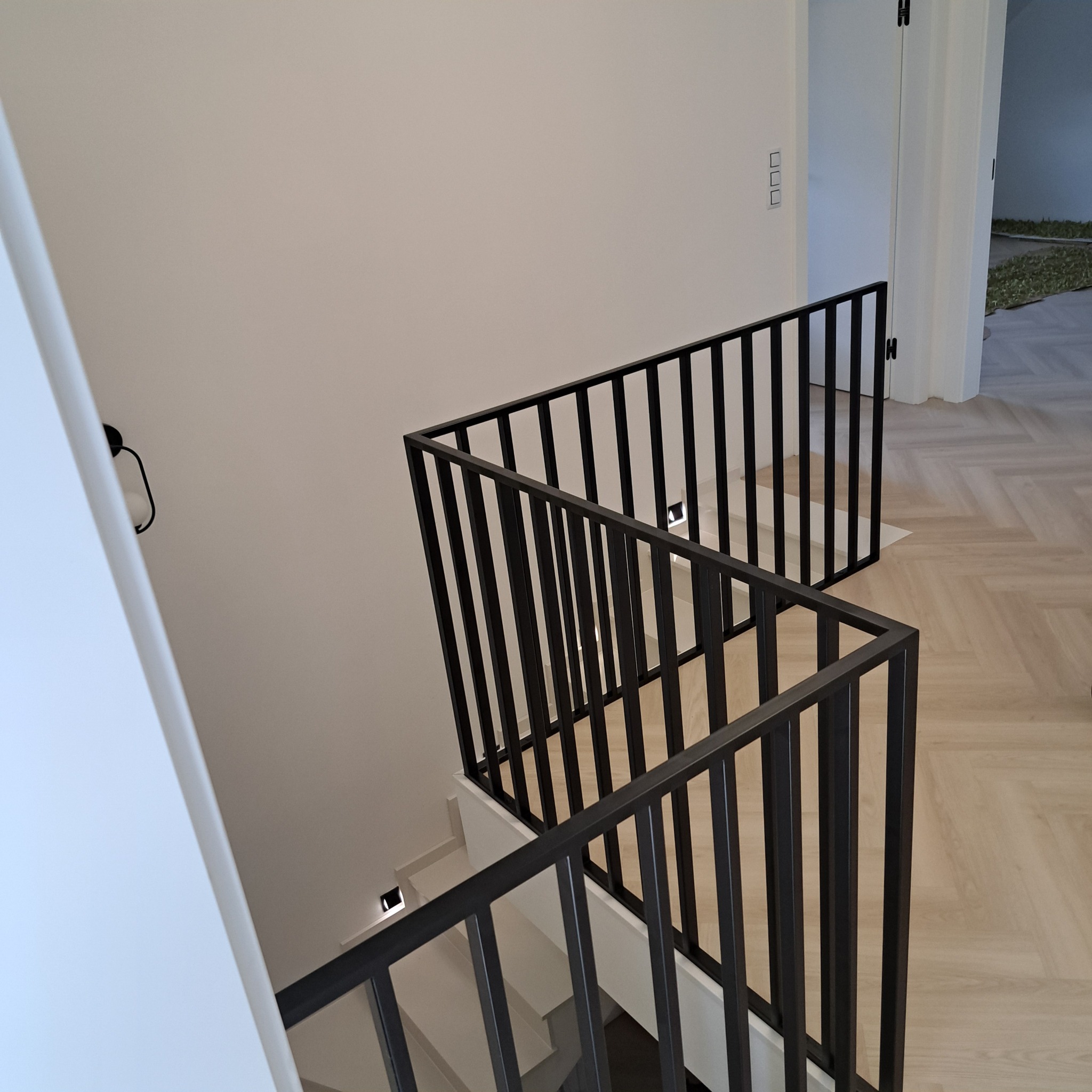 Minimalistyczna, czarna konstrukcja zabezpieczająca schody w nowoczesnym wnętrzu z parkietem ułożonym w jodełkę.