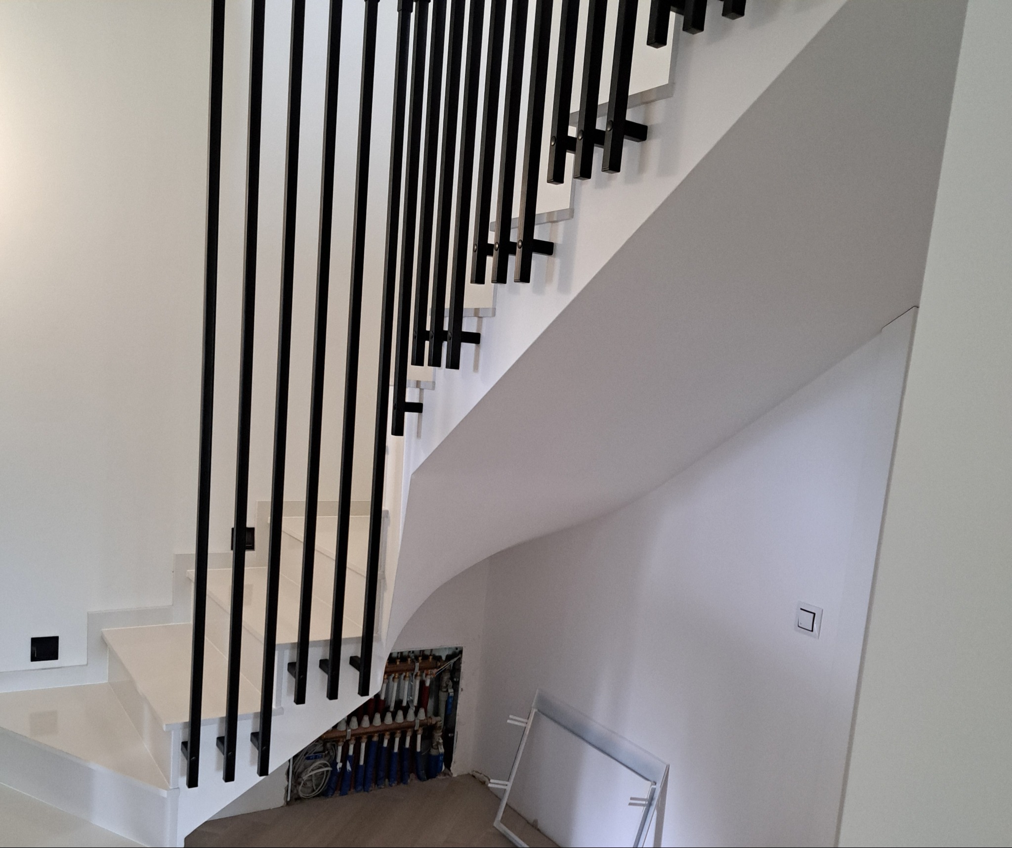 Nowoczesne schody z białymi stopniami i czarnymi, pionowymi elementami zabezpieczającymi, widok z dołu, z ukrytym pod schodami rozdzielaczem instalacji grzewczej.