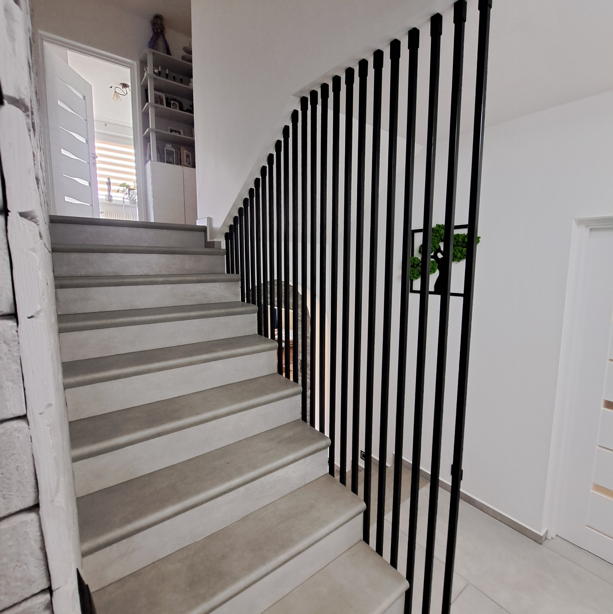 Schody z szarymi stopniami i czarną, pionową konstrukcją zabezpieczającą, ozdobione dekoracją ścienną z mchu w kształcie drzewa.