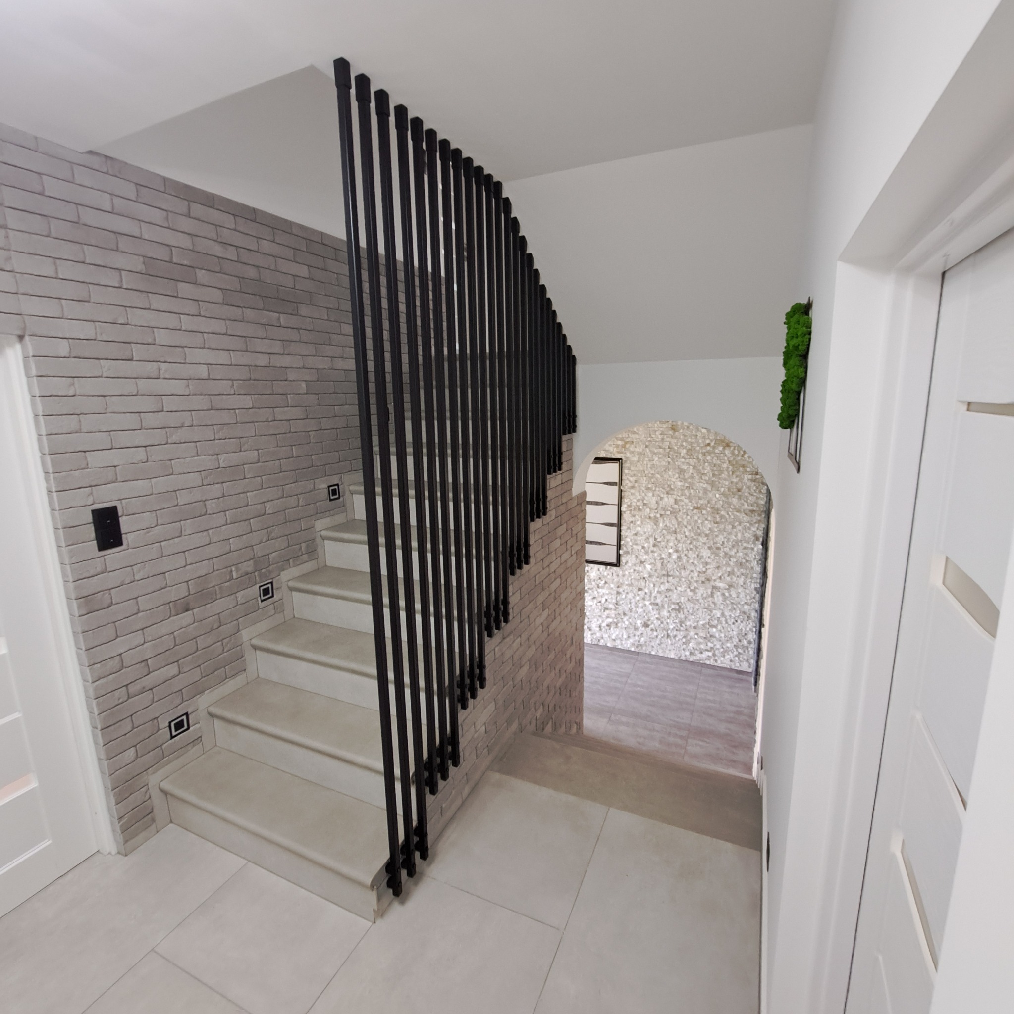Nowoczesna, czarna barierka schodowa z pionowych prętów w domu w Porażu, na tle ściany z cegły i schodów wyłożonych jasnymi płytkami.