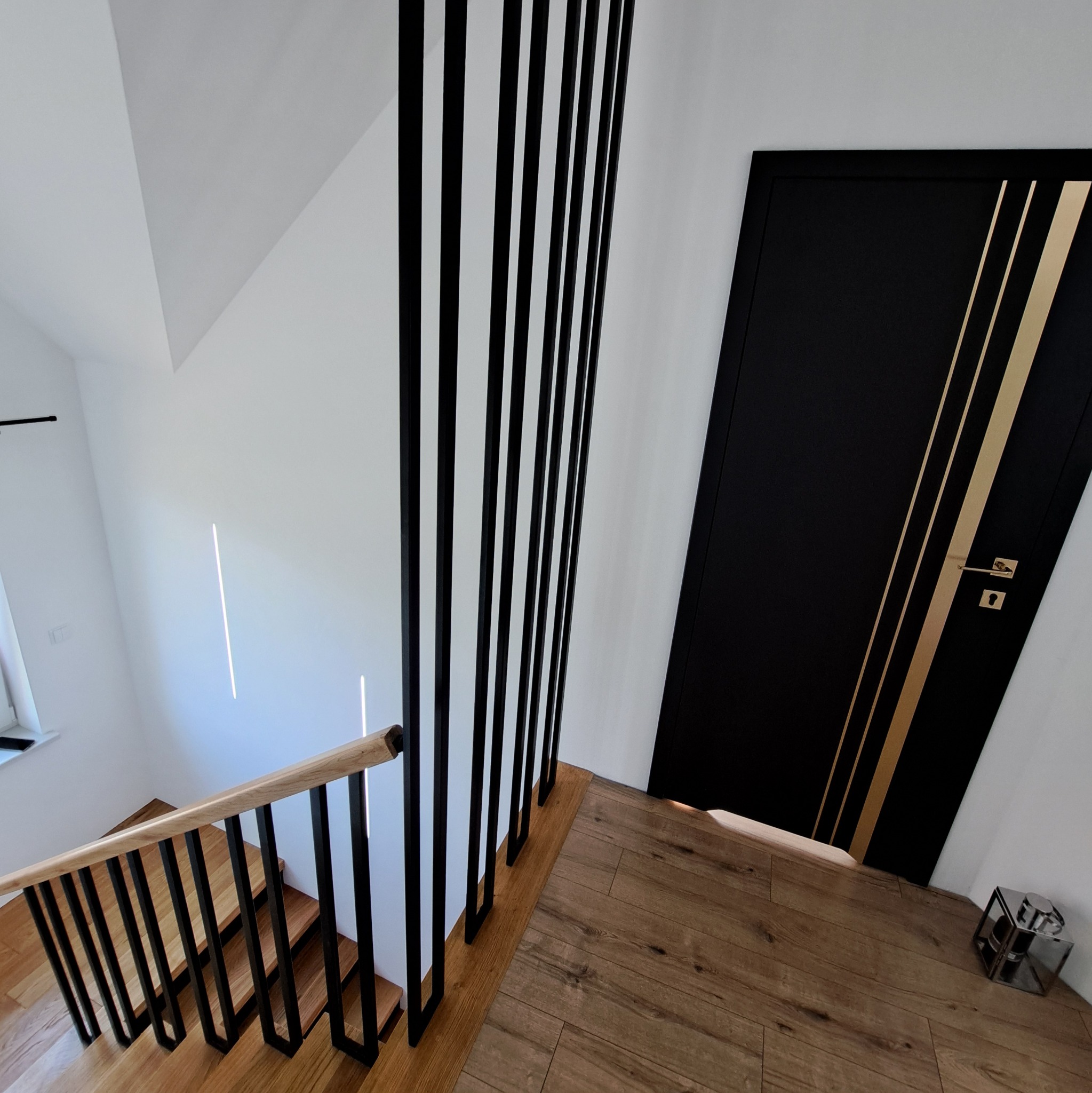 Nowoczesna balustrada schodowa z czarnymi, pionowymi elementami i drewnianą poręczą, widok z góry na schody z drewnianymi stopniami i czarną, stylową drzwią z złotymi paskami w tle.