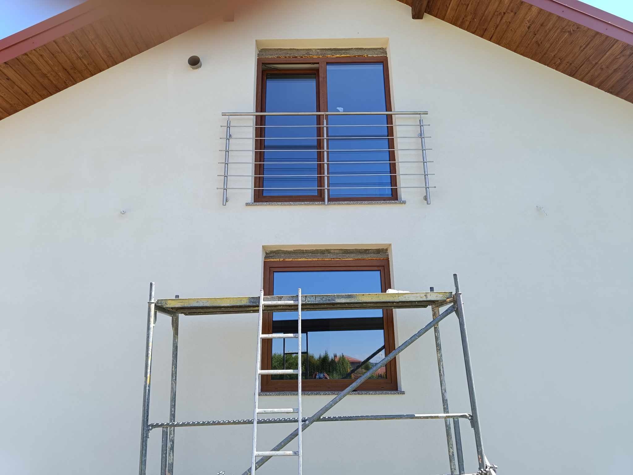Elewacja domu z zamontowaną, poziomą, metalową konstrukcją zabezpieczającą na balkonie oraz rusztowaniem przed oknem na niższej kondygnacji.