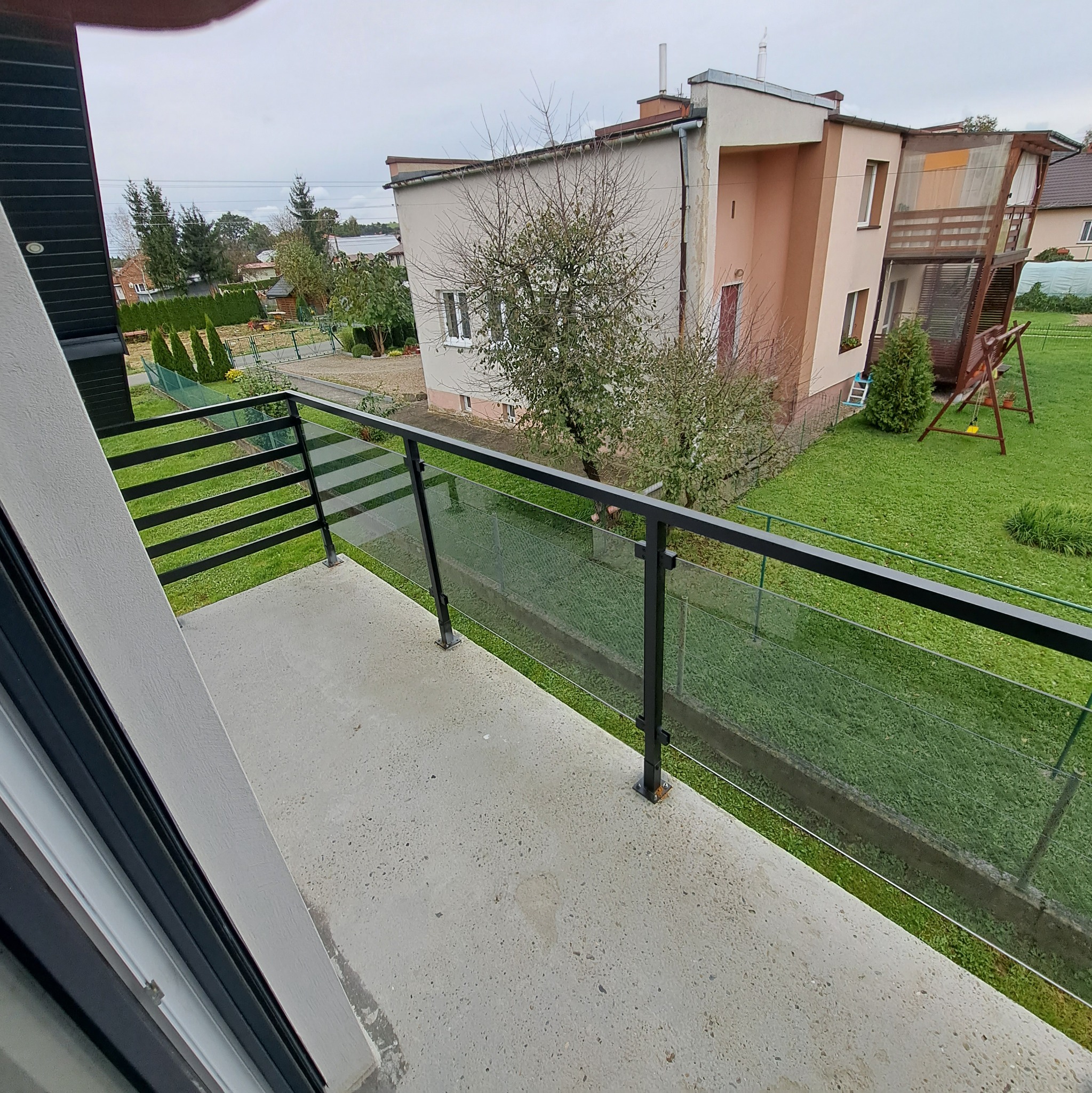 Balkon z czarną ramą i szklanym wypełnieniem, widok na ogród z drzewami i huśtawką, pochmurne niebo.