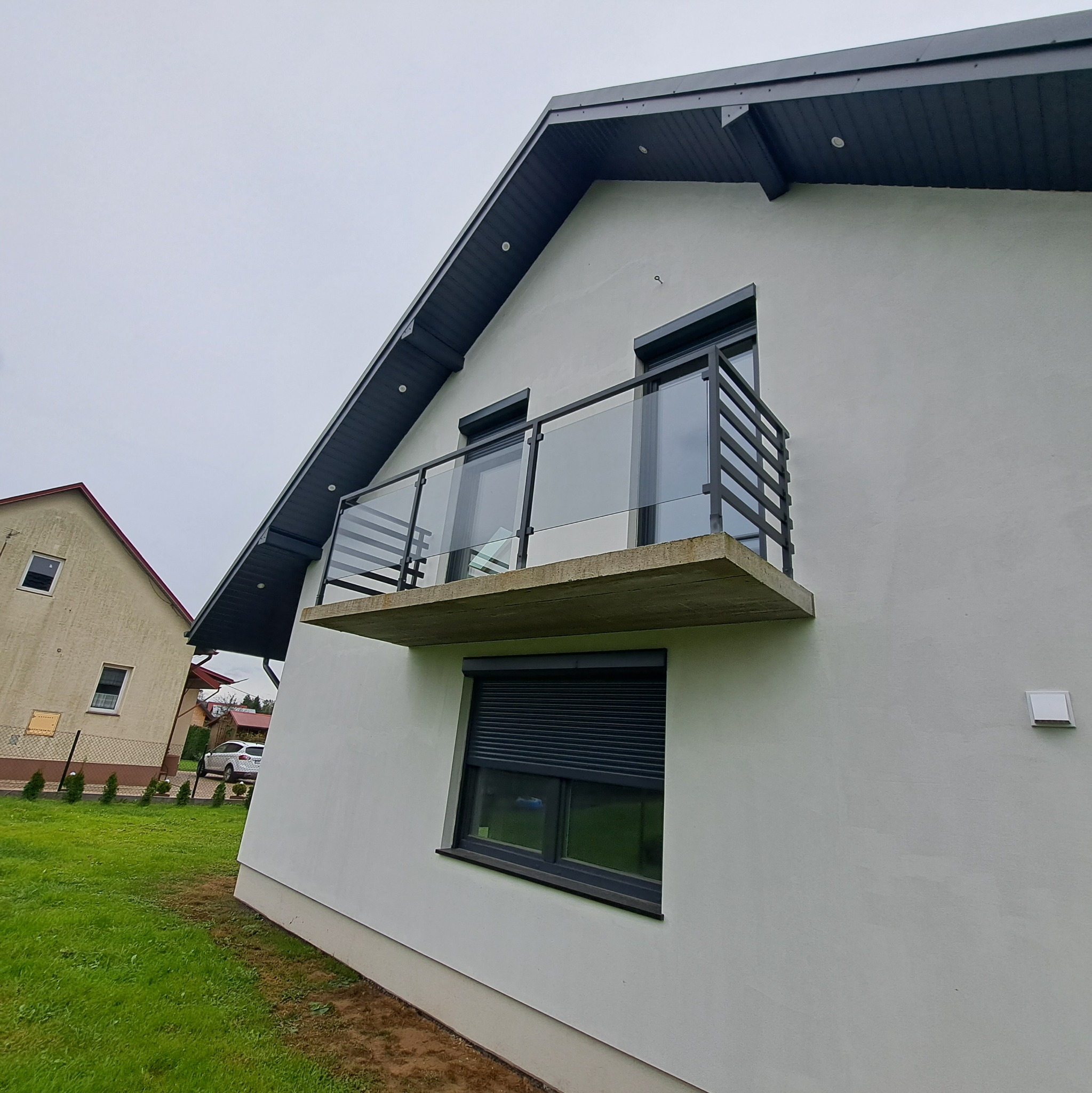 Nowoczesny balkon z czarną metalową konstrukcją i szklanymi panelami, zamontowany na betonowej płycie, widok z dołu na biały budynek z szarym dachem.