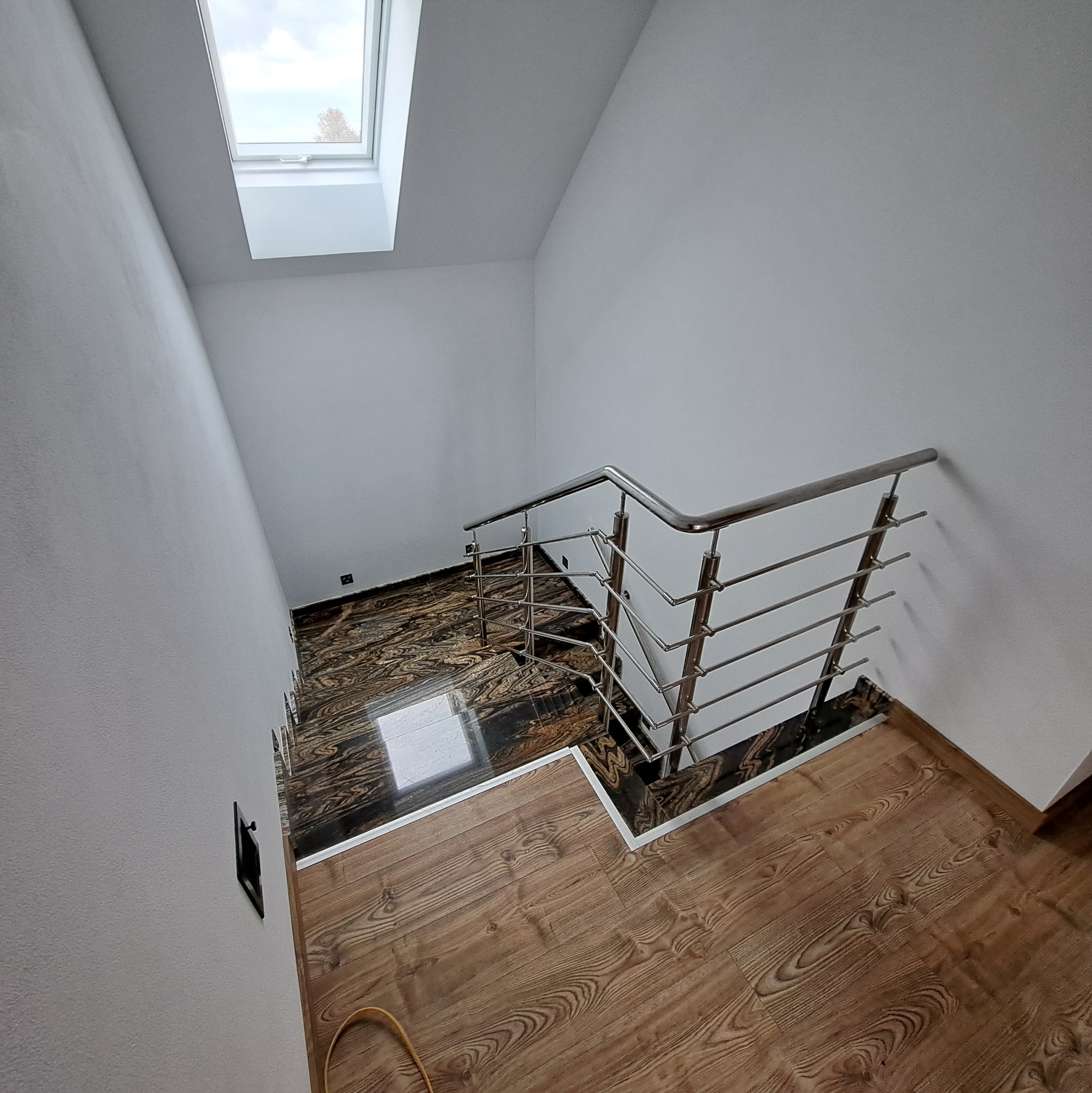 Wewnętrzne schody z drewnopodobnymi panelami i marmurowymi stopniami, zabezpieczone metalową konstrukcją z poziomymi prętami, oświetlone światłem z okna dachowego.